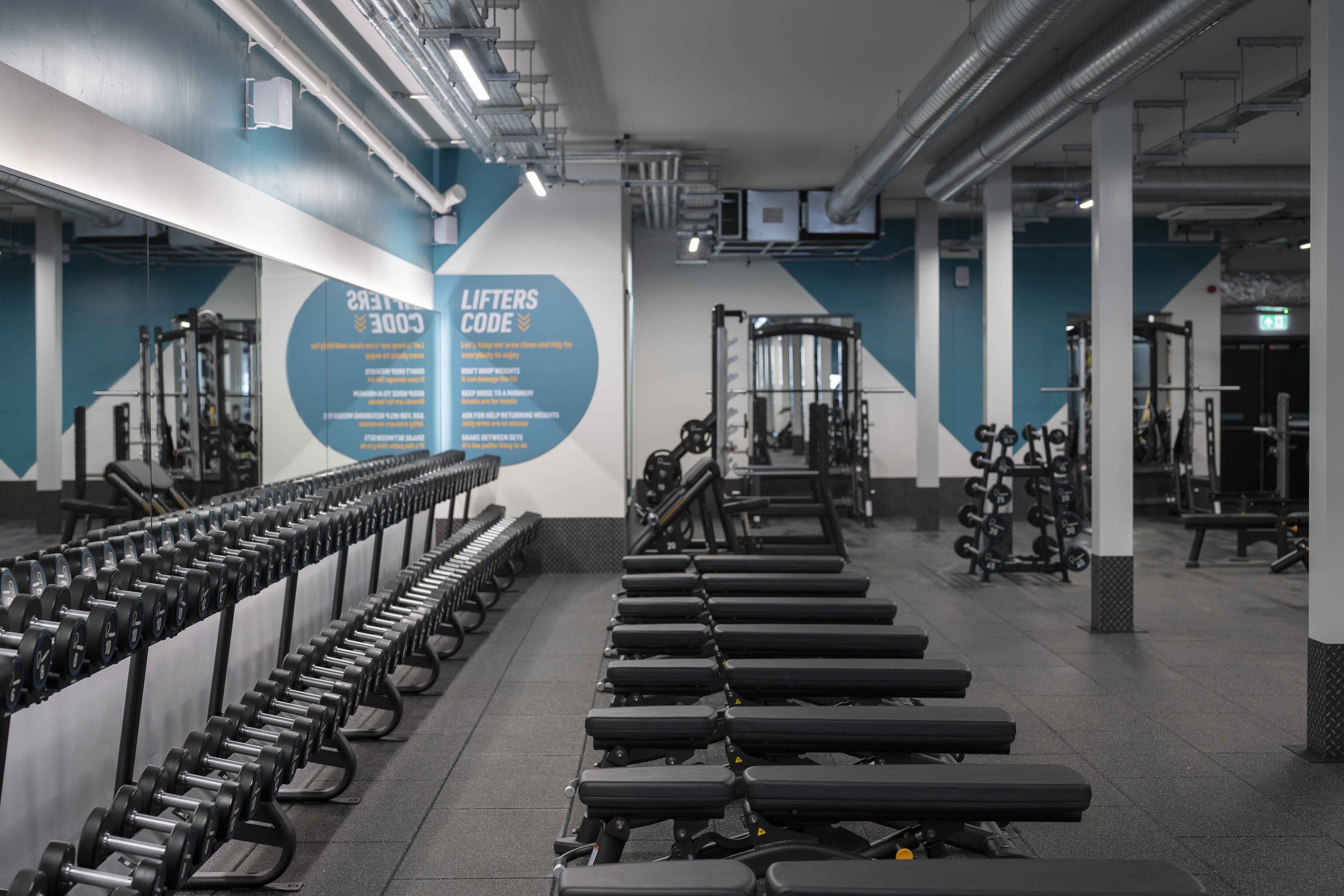 Interior of a gym with free weights, cable machines, workout benches, and a large mirror reflecting the equipment and a wall with signage and blue accents.