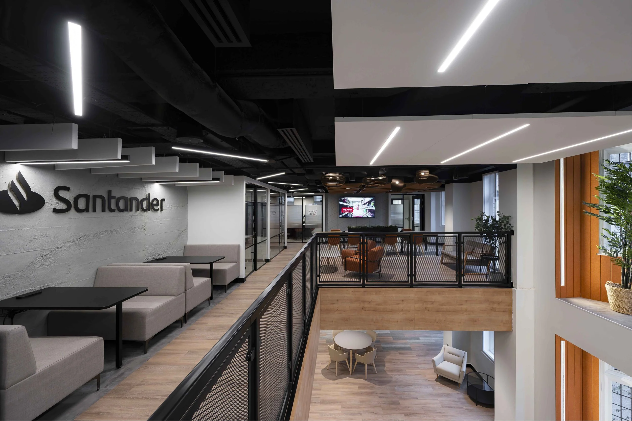Modern office lobby with seating areas, a large wall with logo 'Santander,' and a second-story balcony with seating and decor, illuminated by ceiling lights.