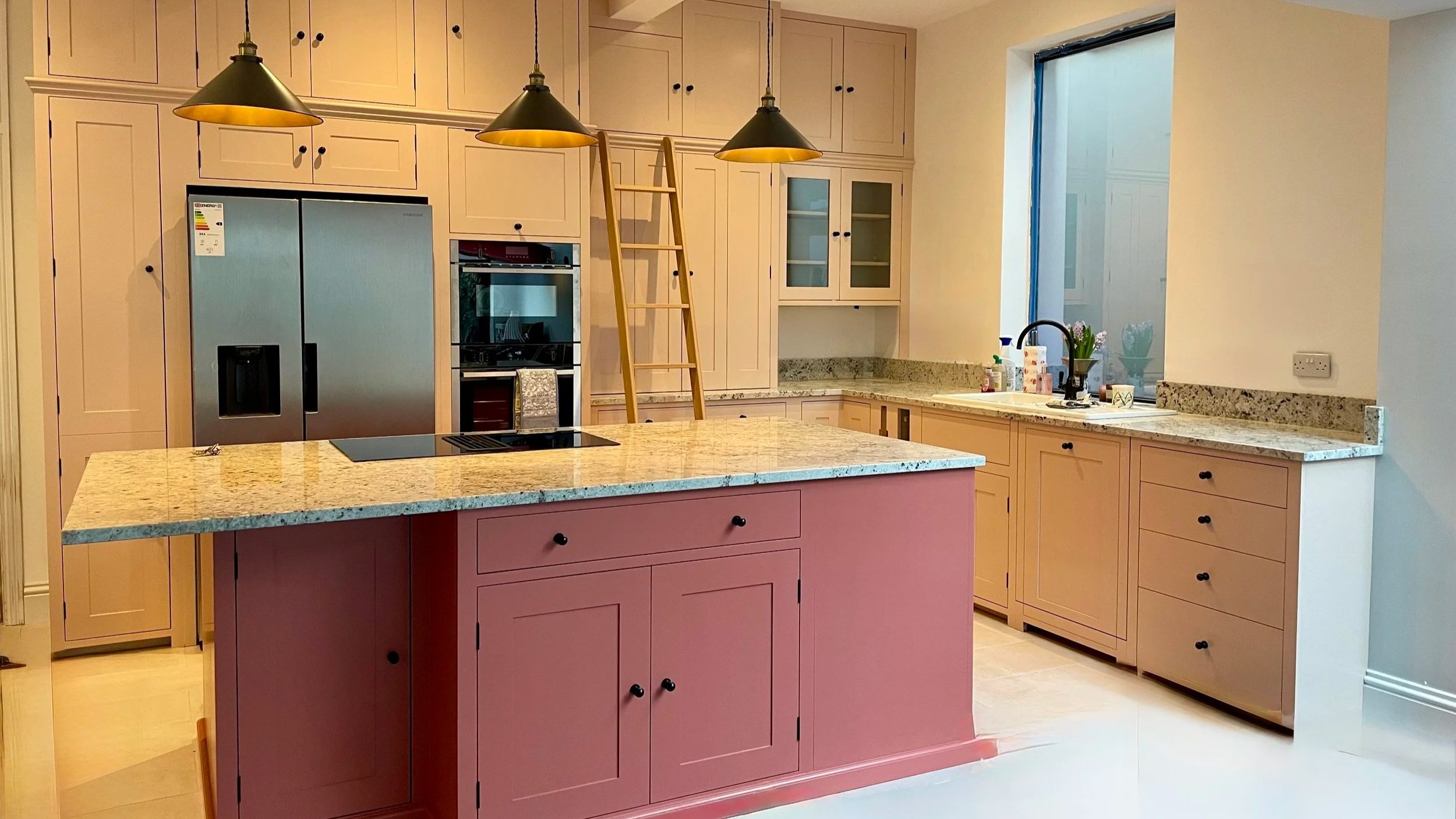 A modern kitchen with light-colored cabinets, granite countertops, and a pink kitchen island with black knobs. There are three pendant lights hanging above the island, a stainless steel refrigerator, oven, built-in microwave, and a wooden ladder lean