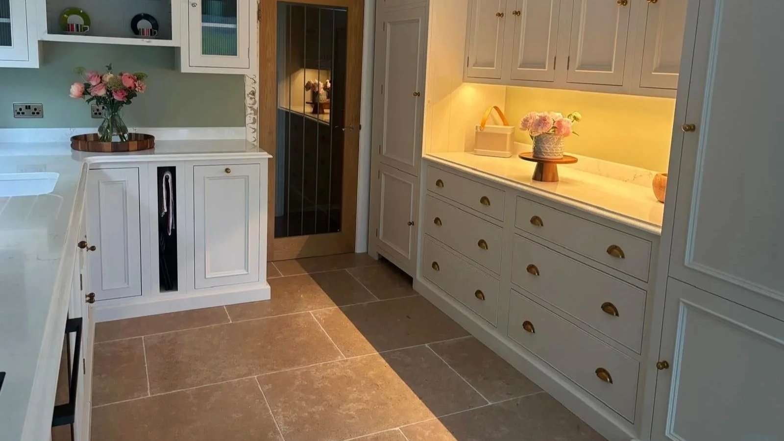 Bespoke fitted kitchen with white cabinetry, brass details and warm under‑cabinet lighting, creating a calm, elegant space with classic charm.