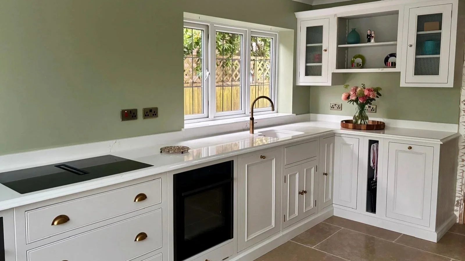 Bespoke fitted kitchen with white cabinetry, brass details and a bright sink area beneath a large window, creating a calm, welcoming cooking space.