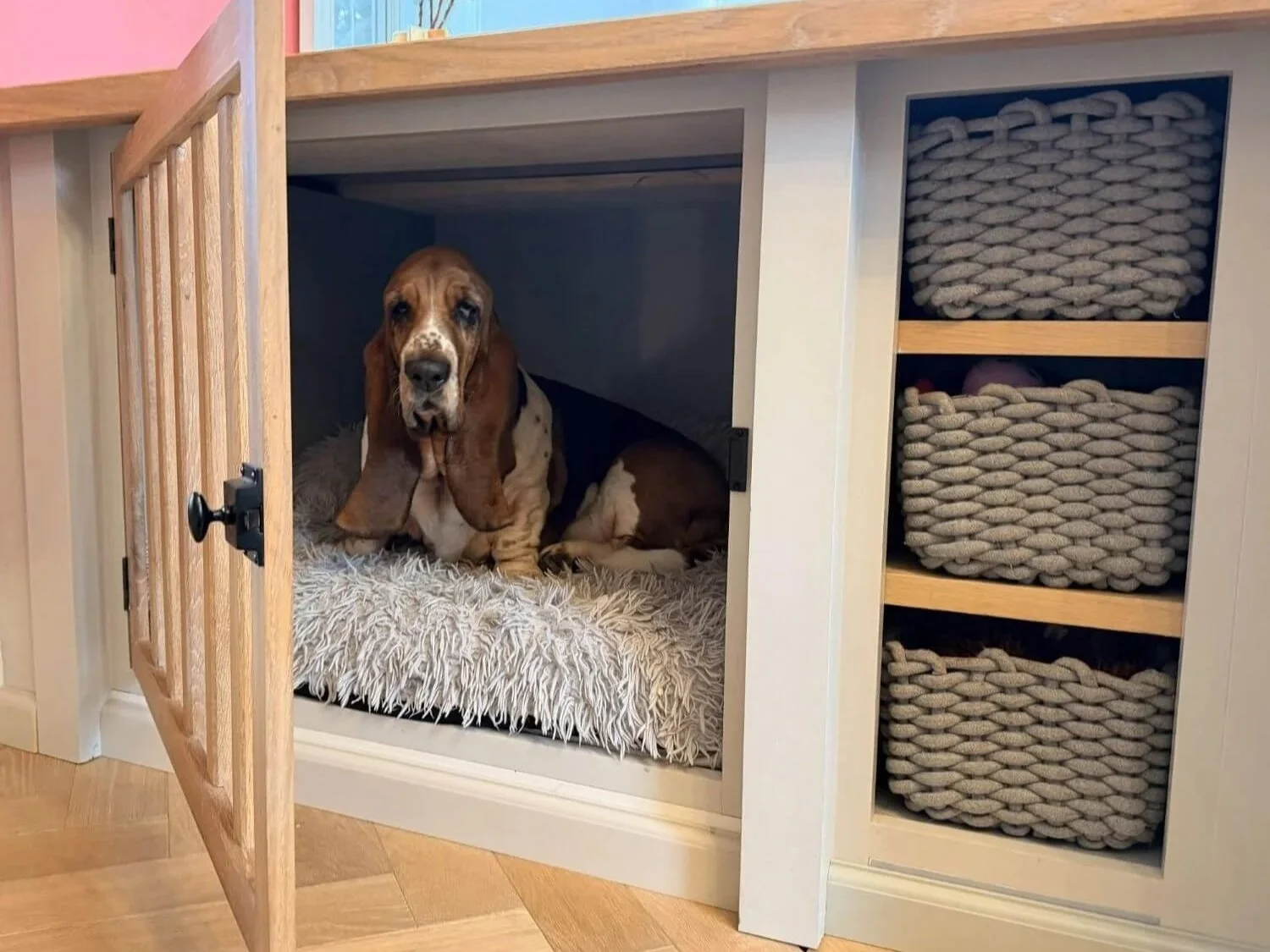 A Basset Hound dog lying inside a wooden dog crate with a fluffy white mat, next to a white cabinet with three woven baskets.