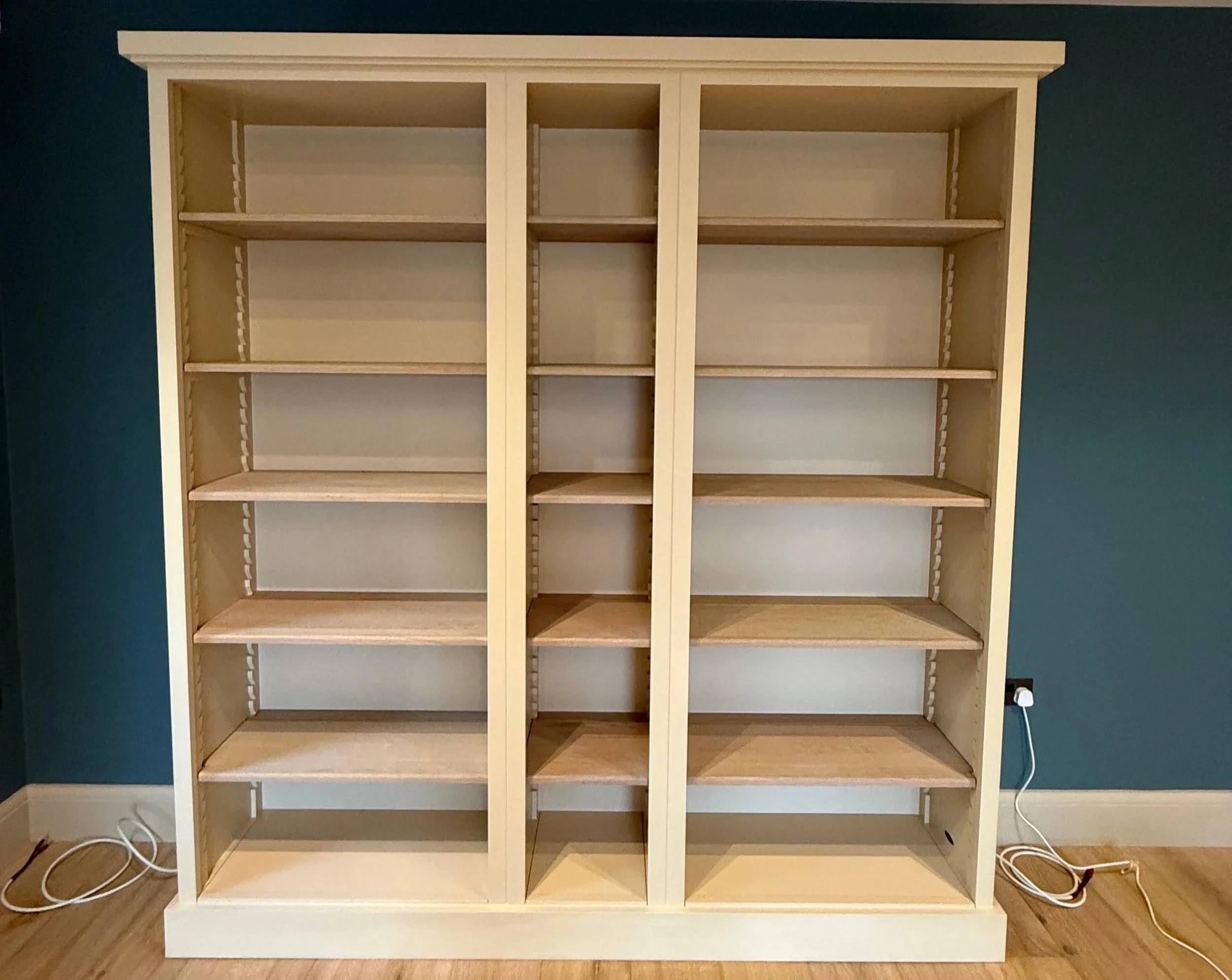 Bespoke built‑in shelving unit with adjustable wooden shelves and a cream frame, handcrafted for flexible, organised storage against a teal wall.