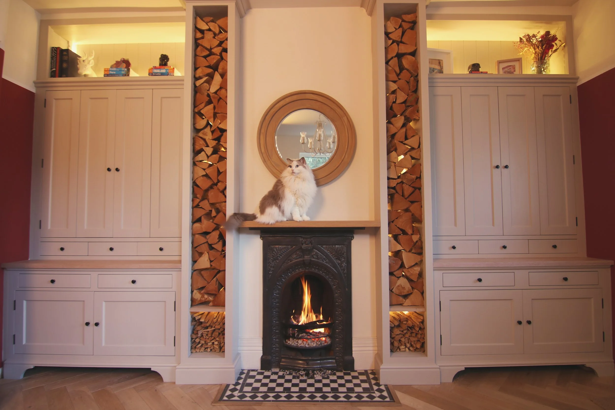 Bespoke living‑room cabinetry with a central cast‑iron fireplace, stacked firewood and warm backlighting, handcrafted to create a cosy, symmetrical focal point.