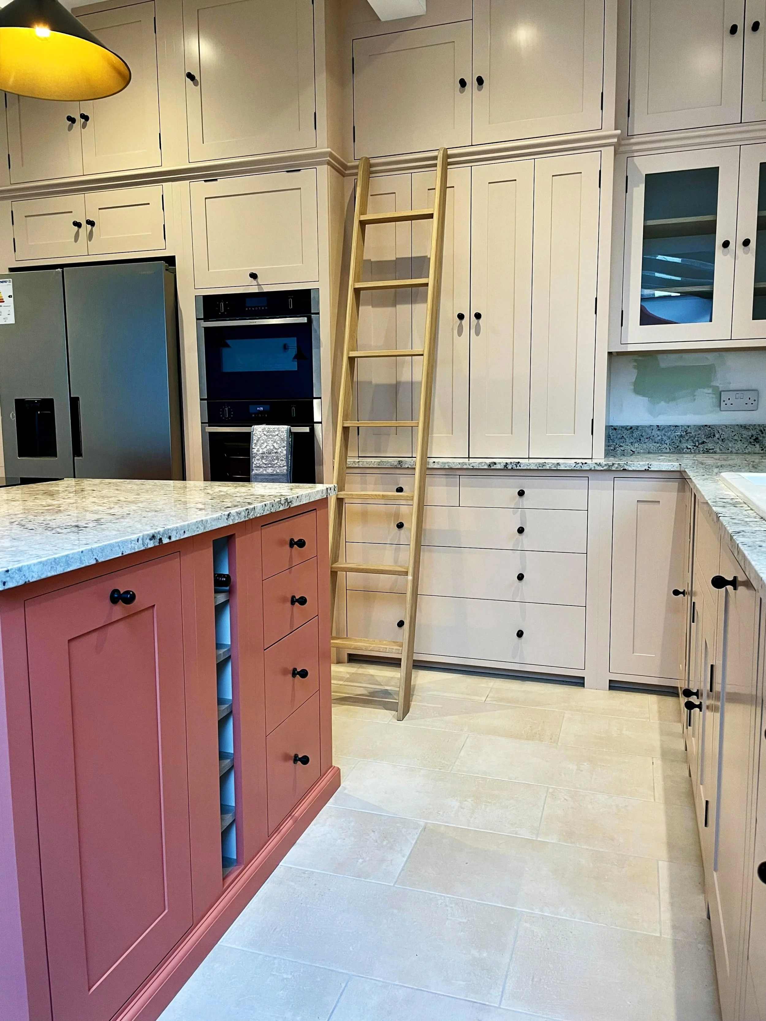 Bespoke fitted kitchen with cream cabinetry, coral island and a tall ladder for high storage, with counter top cabinets to hide appliances. Handcrafted for a warm, functional space in London and South England.