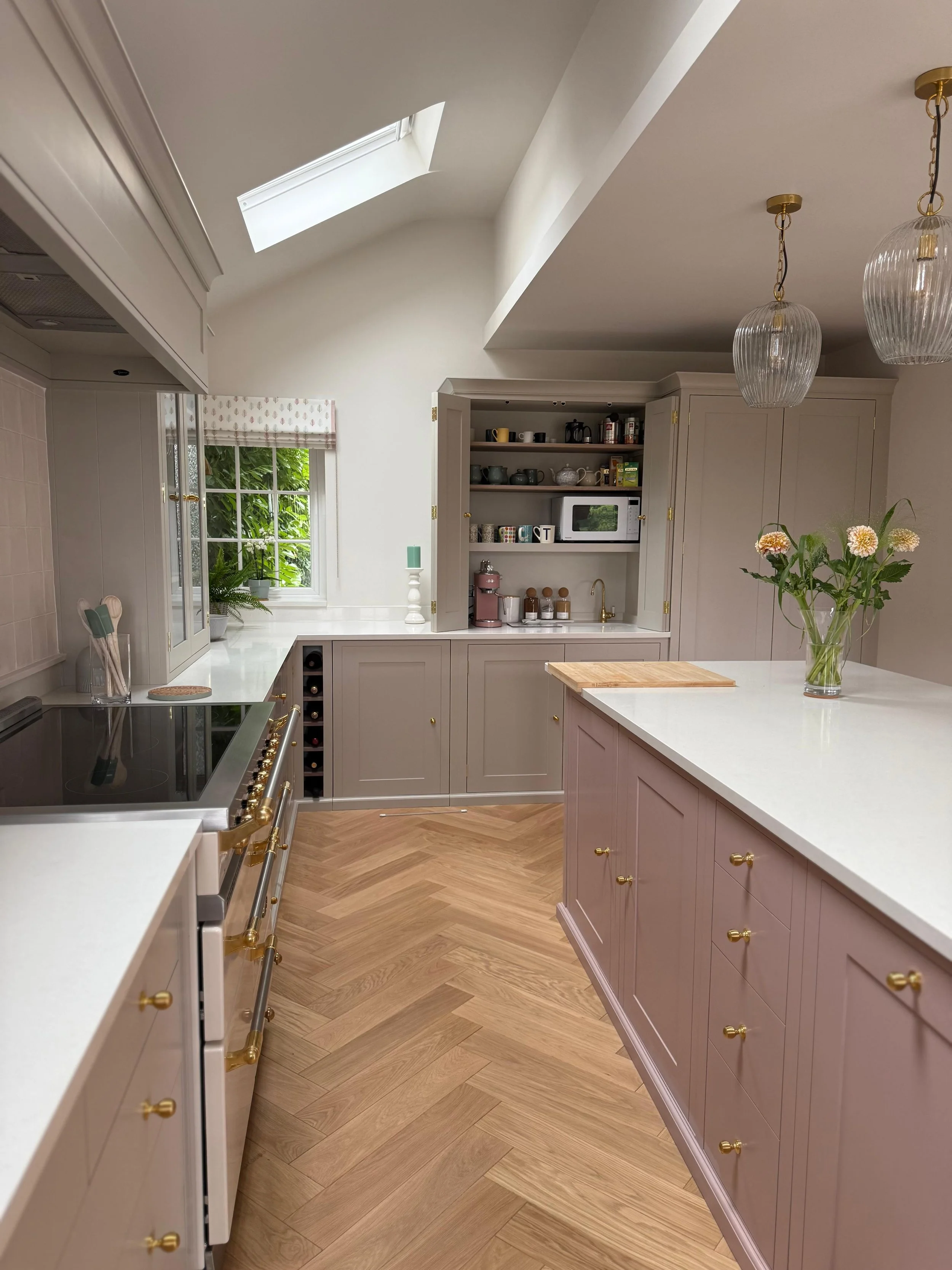 Handmade bespoke kitchen with a pink island, cream cabinetry and gold details, featuring a counter‑top cabinet that neatly hides the kettle, microwave, coffee machine and sink from view.