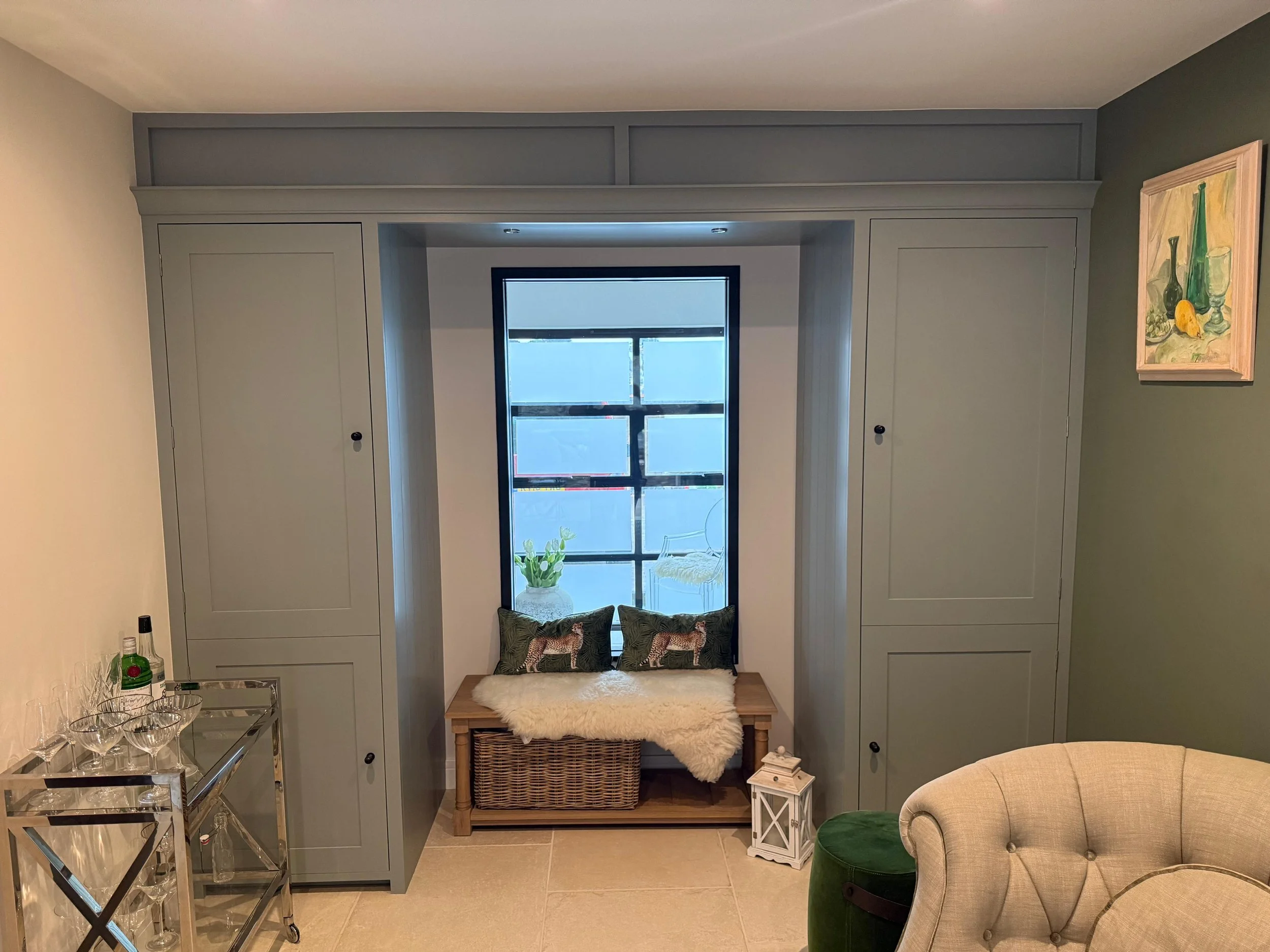 Bespoke built‑in window seat with grey cabinetry, soft cushions and a wicker storage basket, handcrafted to create a cosy, elegant reading nook with natural light.
