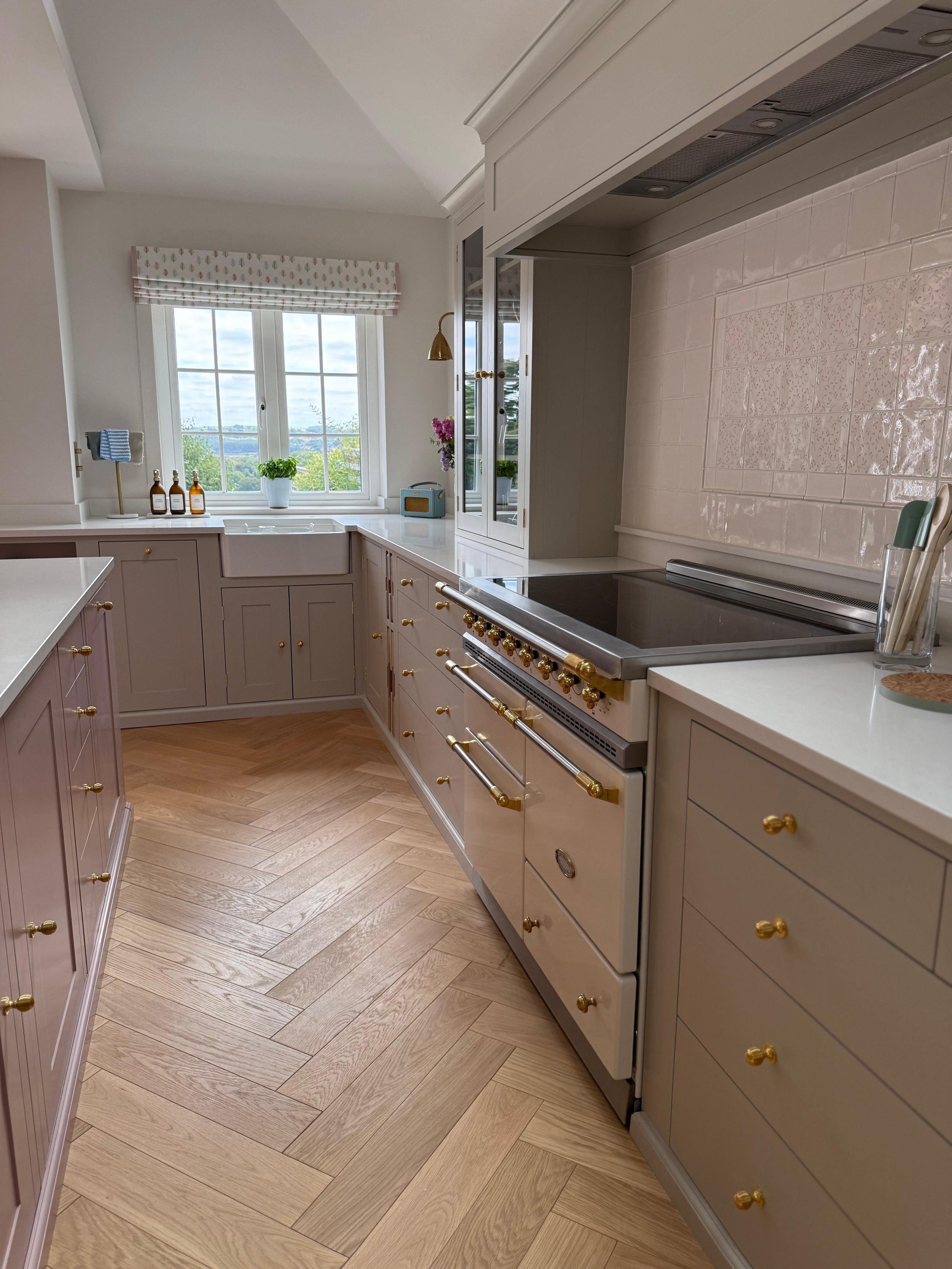 Bespoke fitted kitchen with cream cabinetry, gold details and a white farmhouse sink beneath a window, featuring a pink tiled splashback and a premium range cooker.