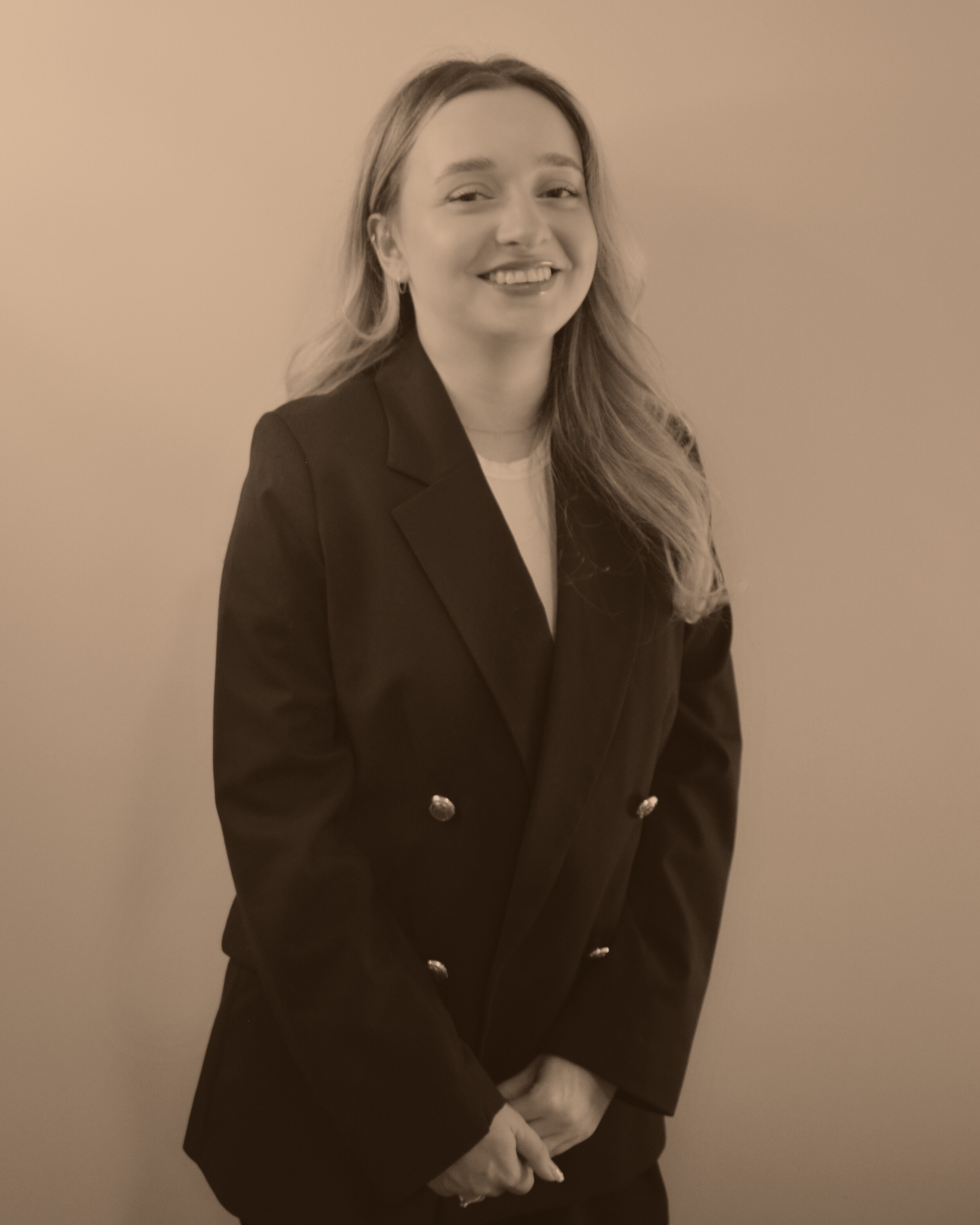 A woman smiling, wearing a dark blazer with metallic buttons, standing against a plain wall.
