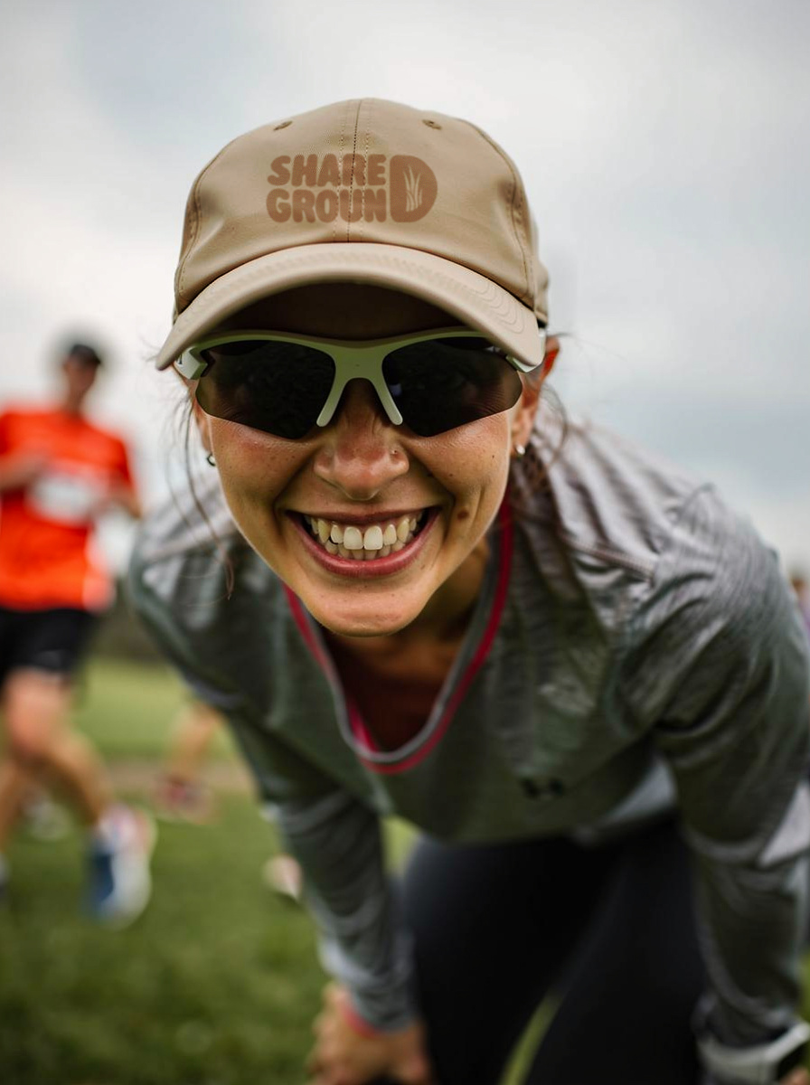 A woman smiling and wearing sunglasses and a beige cap with 'Share Ground' logo, participating in an outdoor athletic event with other runners.