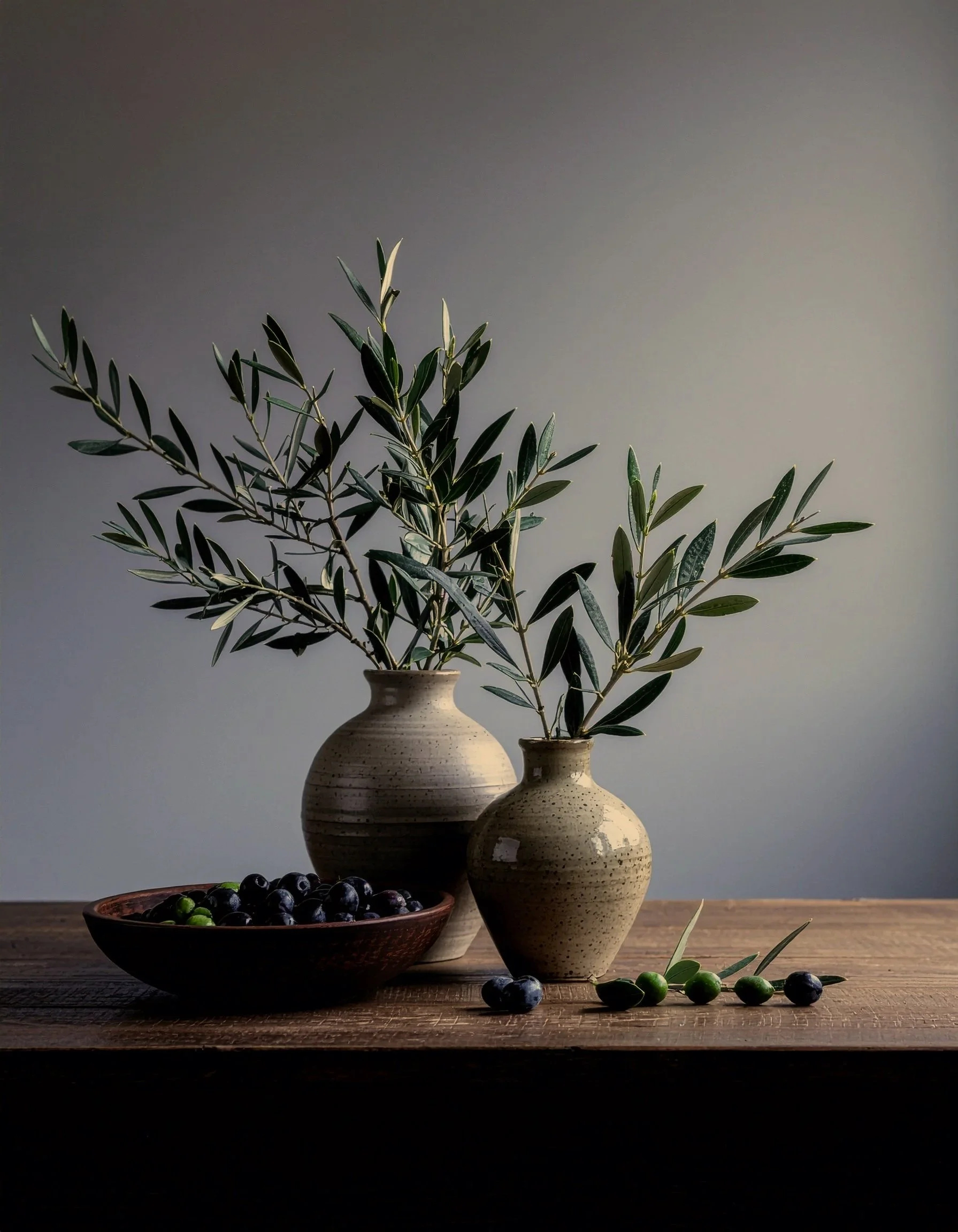 Still life of two ceramic vases with olive branches, a bowl of blueberries, and some scattered blueberries and olives on a wooden surface with a plain background.