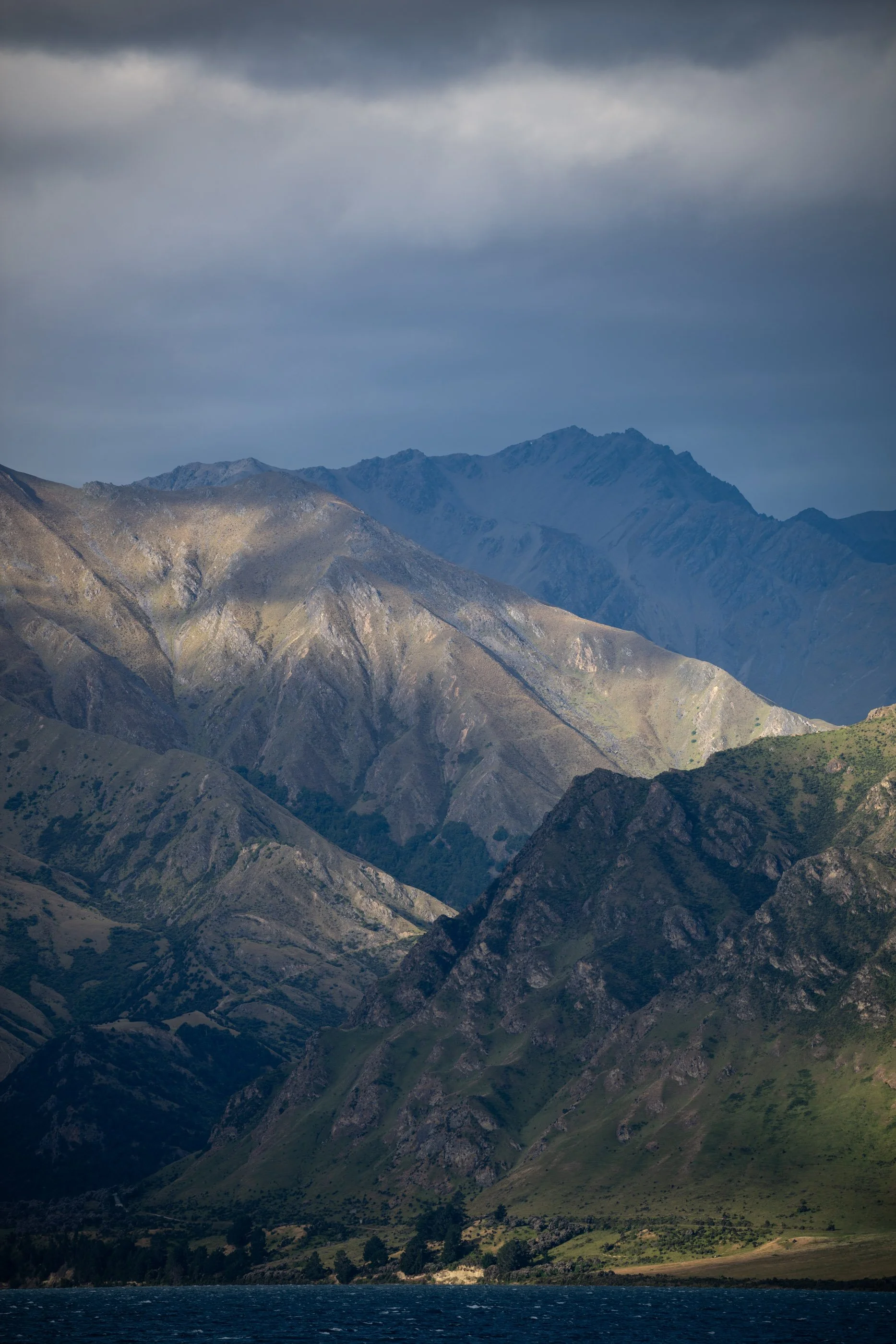 Berglandschaft mit mehreren Bergen und einem See im Vordergrund, bewölkter Himmel.