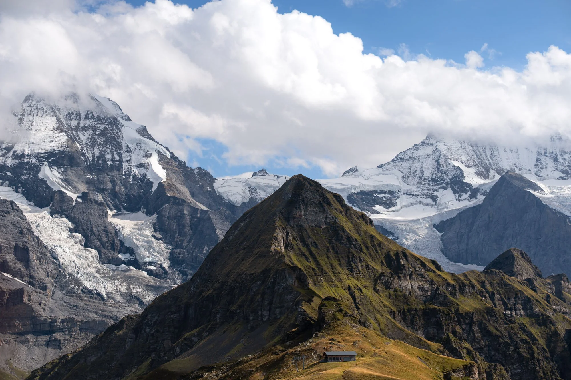 Berglandschaft mit hohen, schneebedeckten Gipfeln, Wolken und einer kleinen Hütte am Fuße des Hügels.