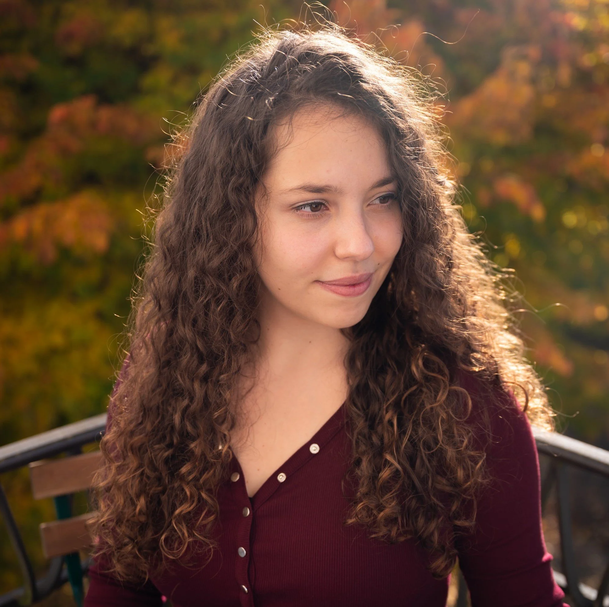 Junge Frau mit lang, lockigem Haar, in einer Herbstlandschaft auf einer Parkbank sitzend. Dalia Schär.
