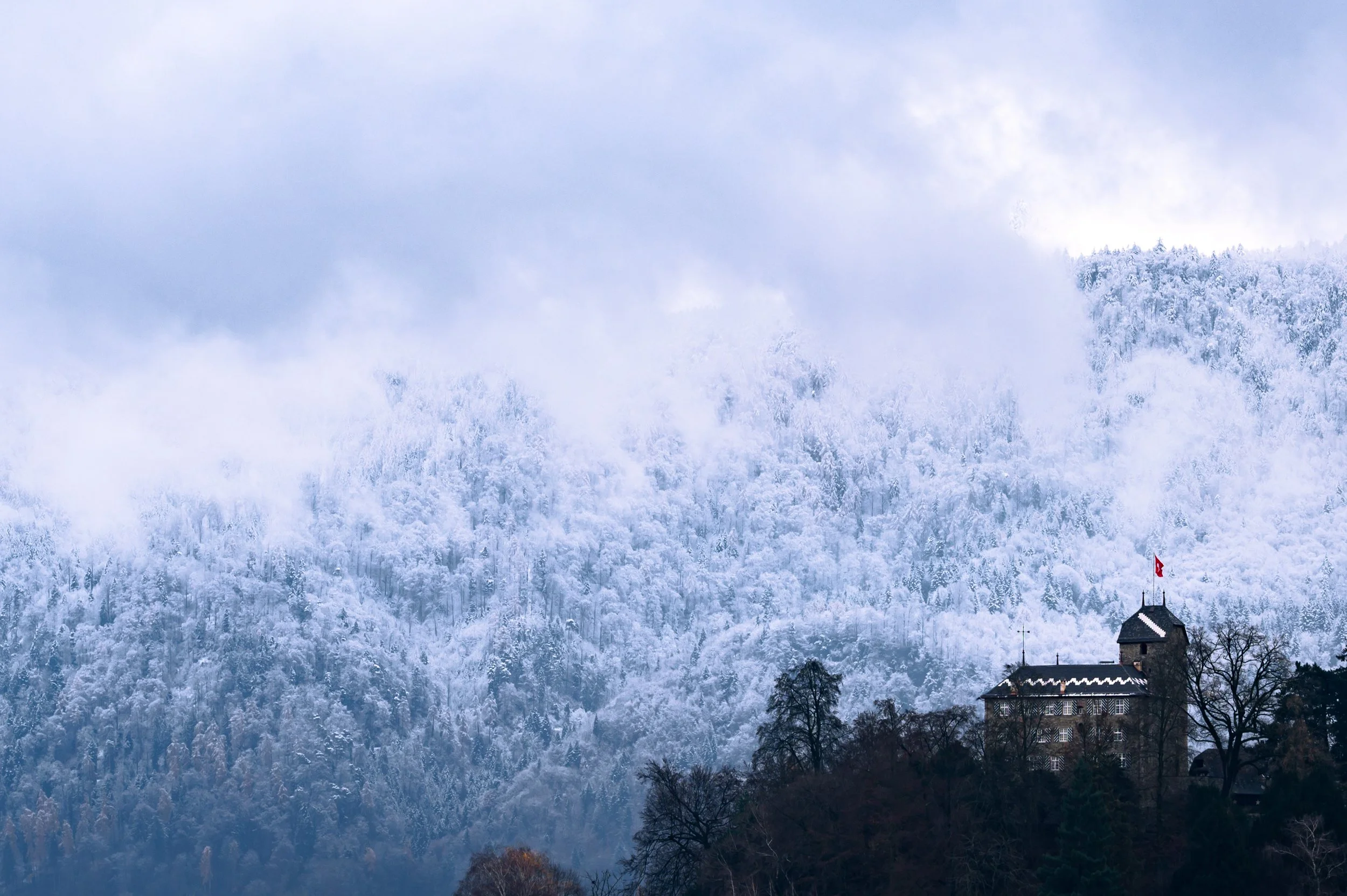 Schneebedeuter Berg im Nebel mit der kleinen Burg von Buonas im Kanton Zug und Schweizer Flagge im Vordergrund.
