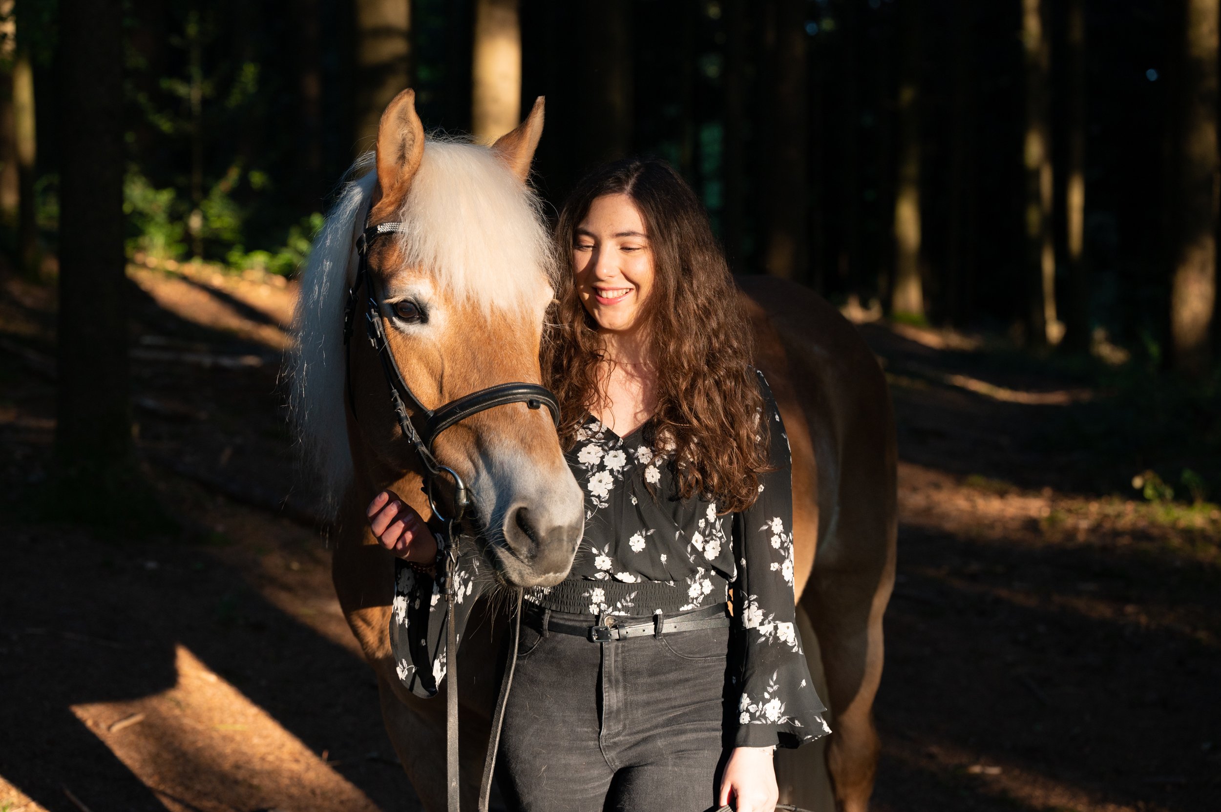 Junge Frau mit braunen, lockigen Haaren, trägt schwarze Hose und eine schwarze Bluse mit weißen Blumen, steht im Wald neben einem braunen Pferd mit weißer Mähne, beide lächeln und schauen sich an.
