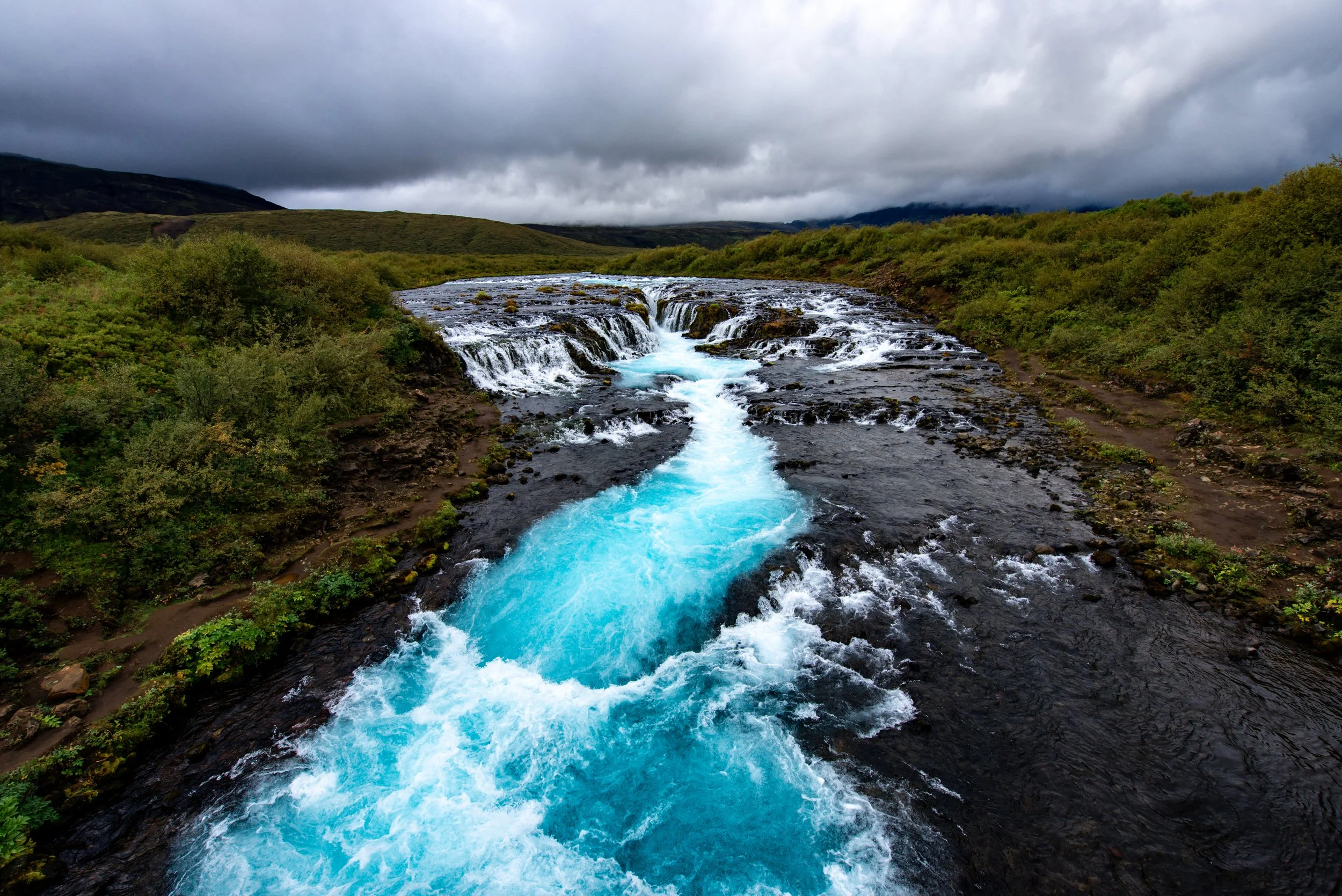 Ein Fluss mit türkisem Wasser fließt über kleine Wasserfälle durch eine grüne Landschaft in Island unter einem bewölkten Himmel.