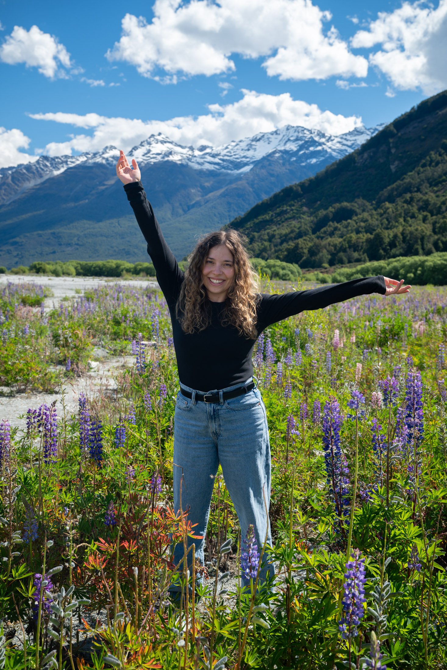Eine lachende junge Frau steht in einem blühenden Blumenfeld mit Bergen im Hintergrund, bei sonnigem Wetter.