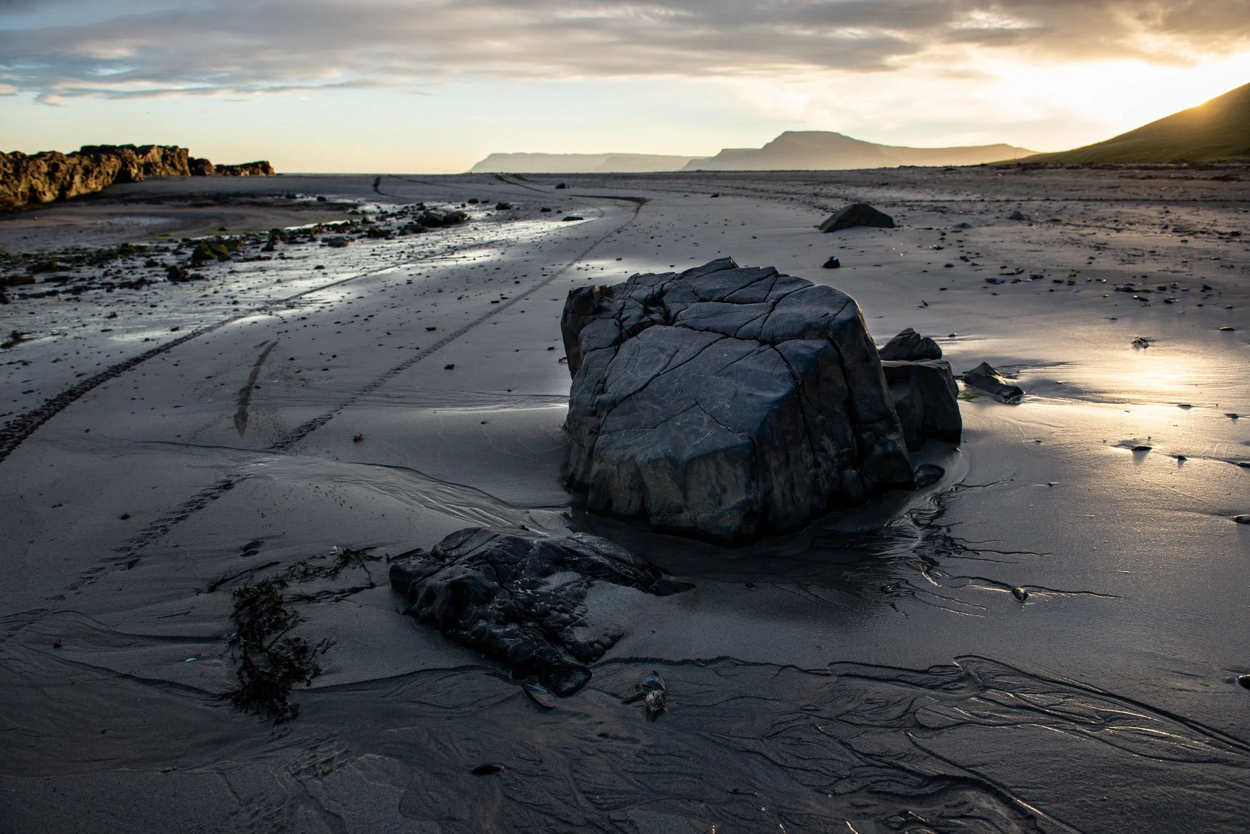 Felsige Küste bei Sonnenuntergang mit dunklem Sandstrand und Spuren im Sand.