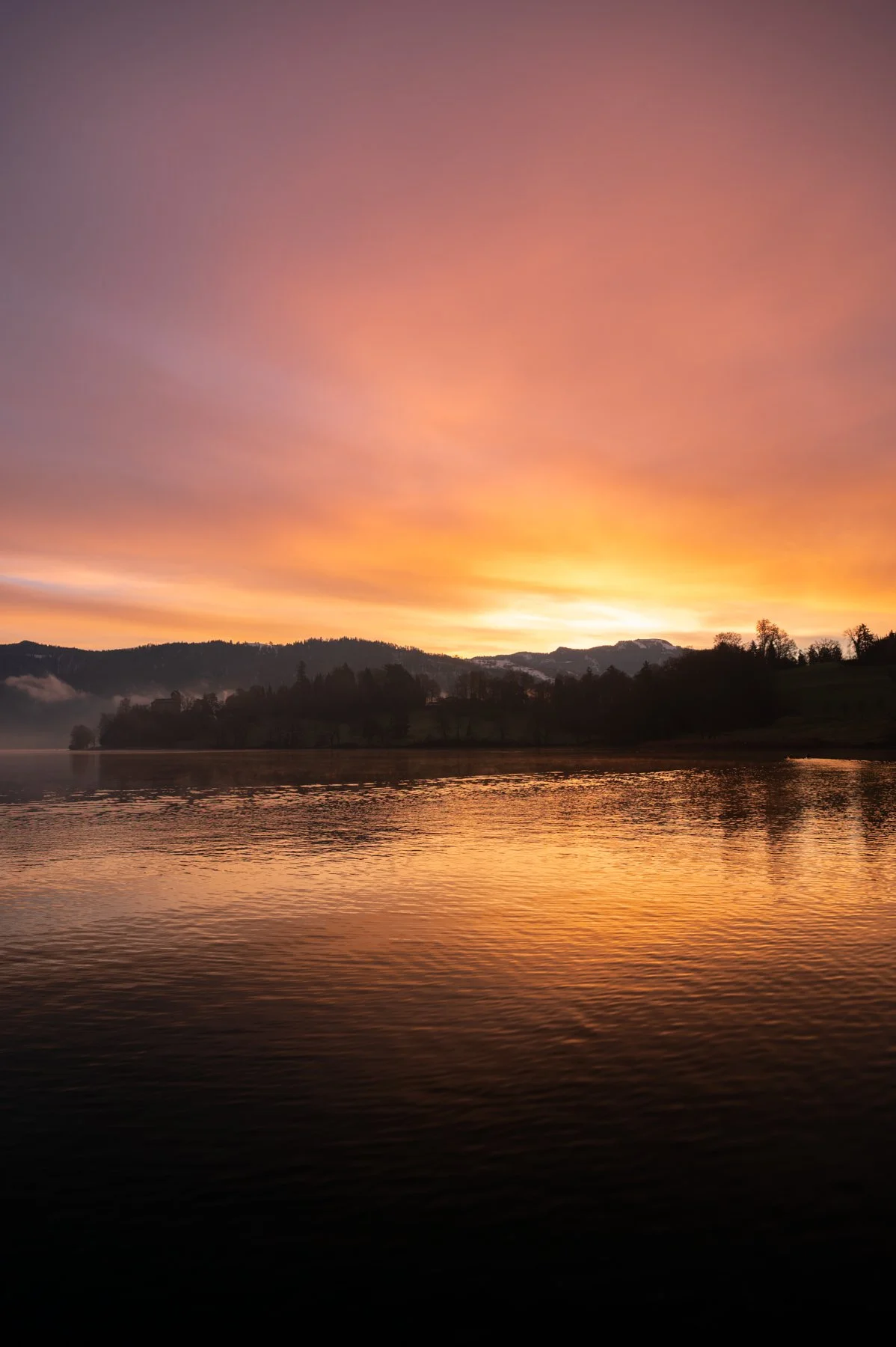 Ein ruhiger Fluss bei Sonnenuntergang mit farbenprächtigem Himmel in Orange, Pink und Lila, umgeben von Hügeln und Bäumen.