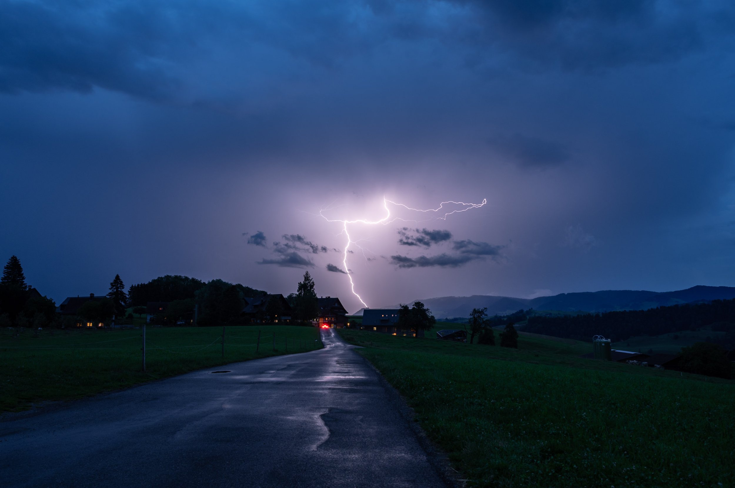 Blitz, der den Himmel bei Nacht erleuchtet, über einer ländlichen Straße mit Häusern und Wiesen, dunkle Wolken am Himmel
