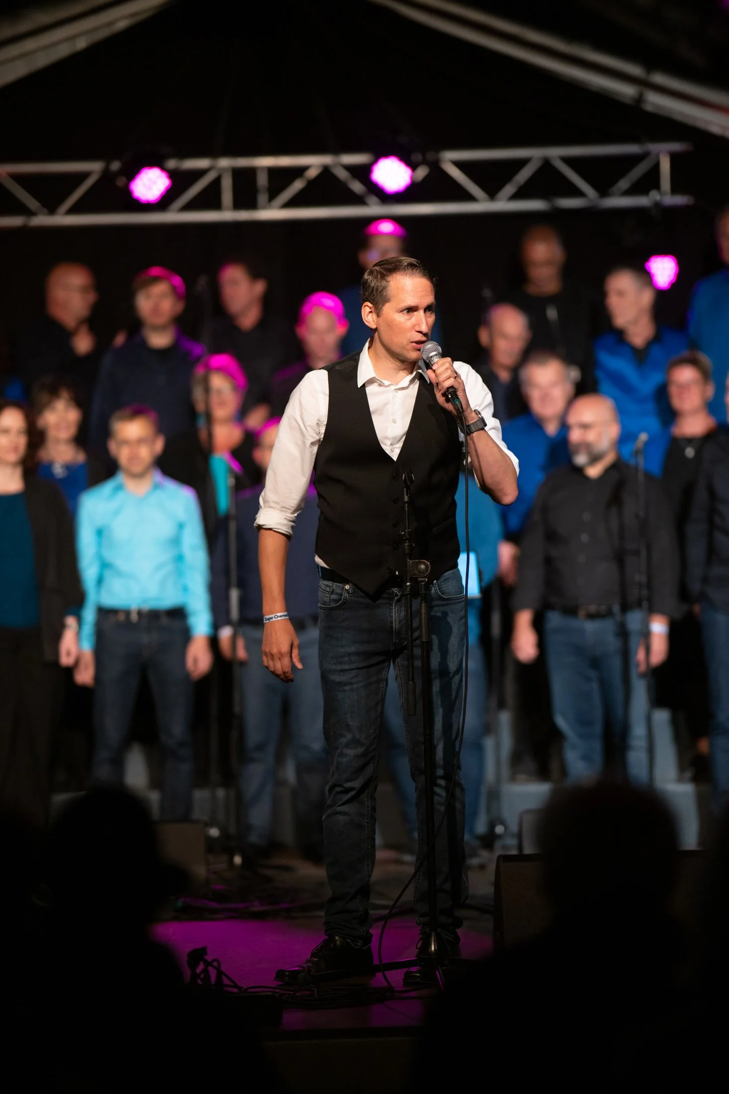 Ein Mann steht mit einem Mikrofon vor einem Chor auf einer Bühne. Im Hintergrund sind viele singende Personen, einige tragen blaue und schwarze Kleidung. Pinke Bühnenlichter leuchten.