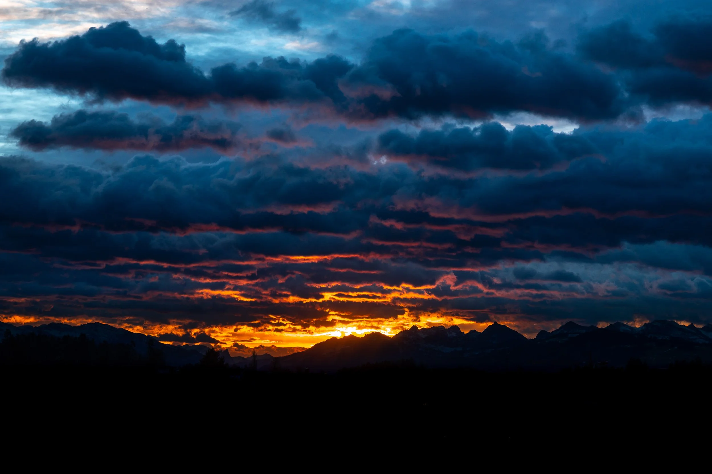 Ein Sonnenuntergang hinter Bergen mit dunklen Wolken am Himmel und orangefarbenem Licht am Horizont.