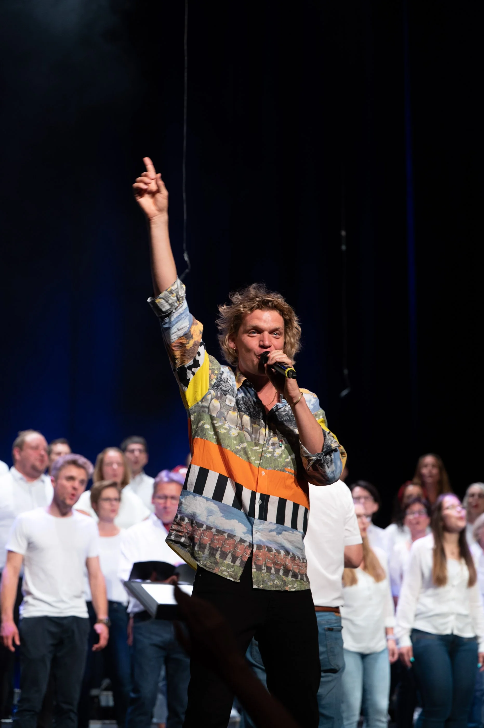 Ein Mann singt in ein Mikrofon, umgeben von einem Chor auf einer Bühne während eines Live-Auftritts.