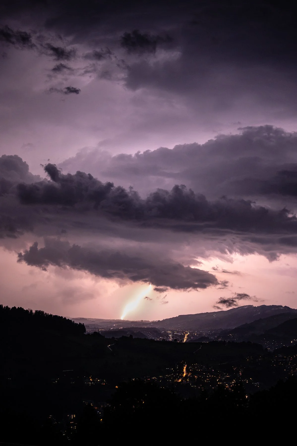 Blitzschlag während eines Gewitters über einer bergigen Stadtlandschaft bei Nacht mit dunklen Wolken.