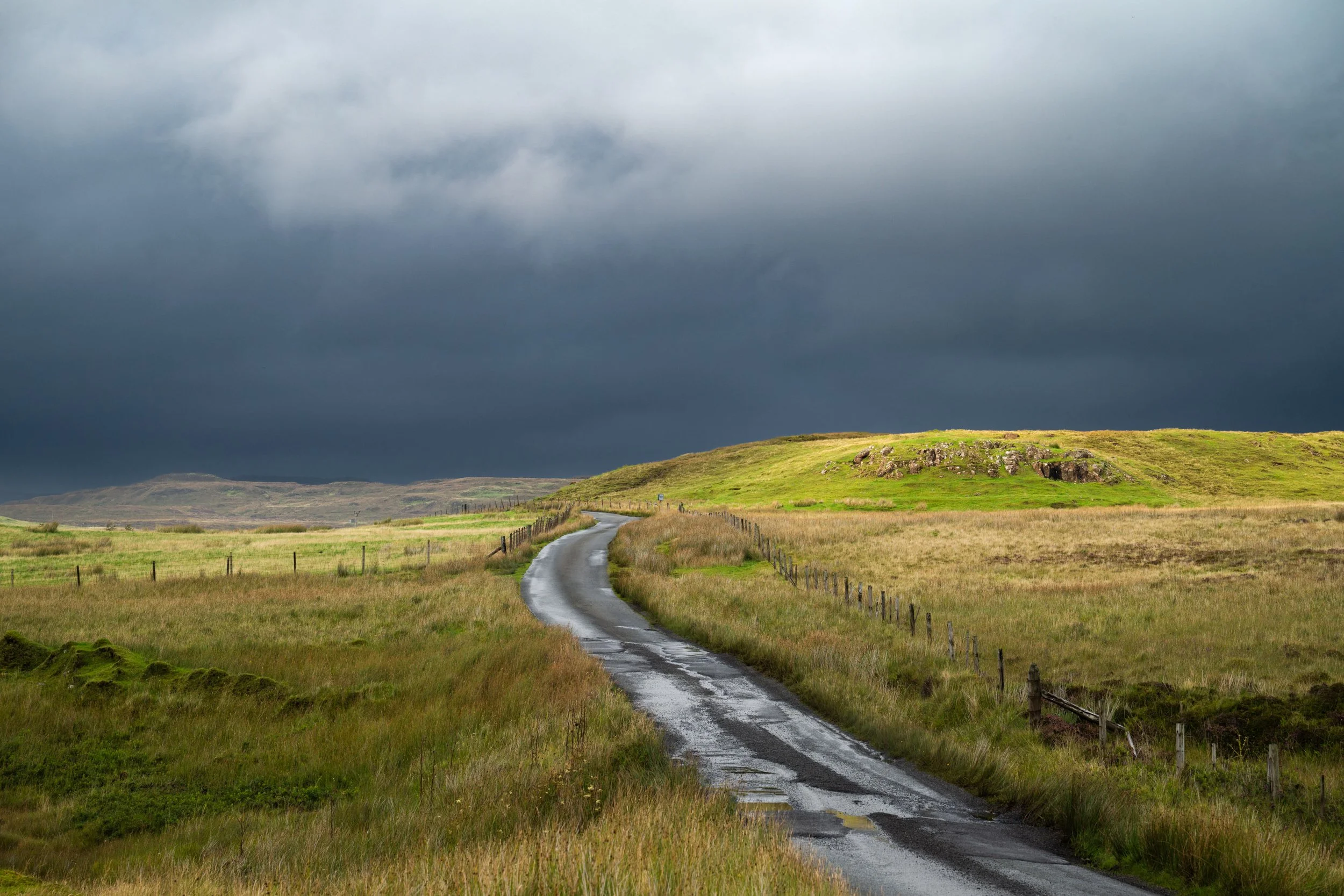 Ein schmaler, nasser Weg führt durch eine grüne Landschaft mit Hügeln; dunkle Wolken hängen am Himmel.