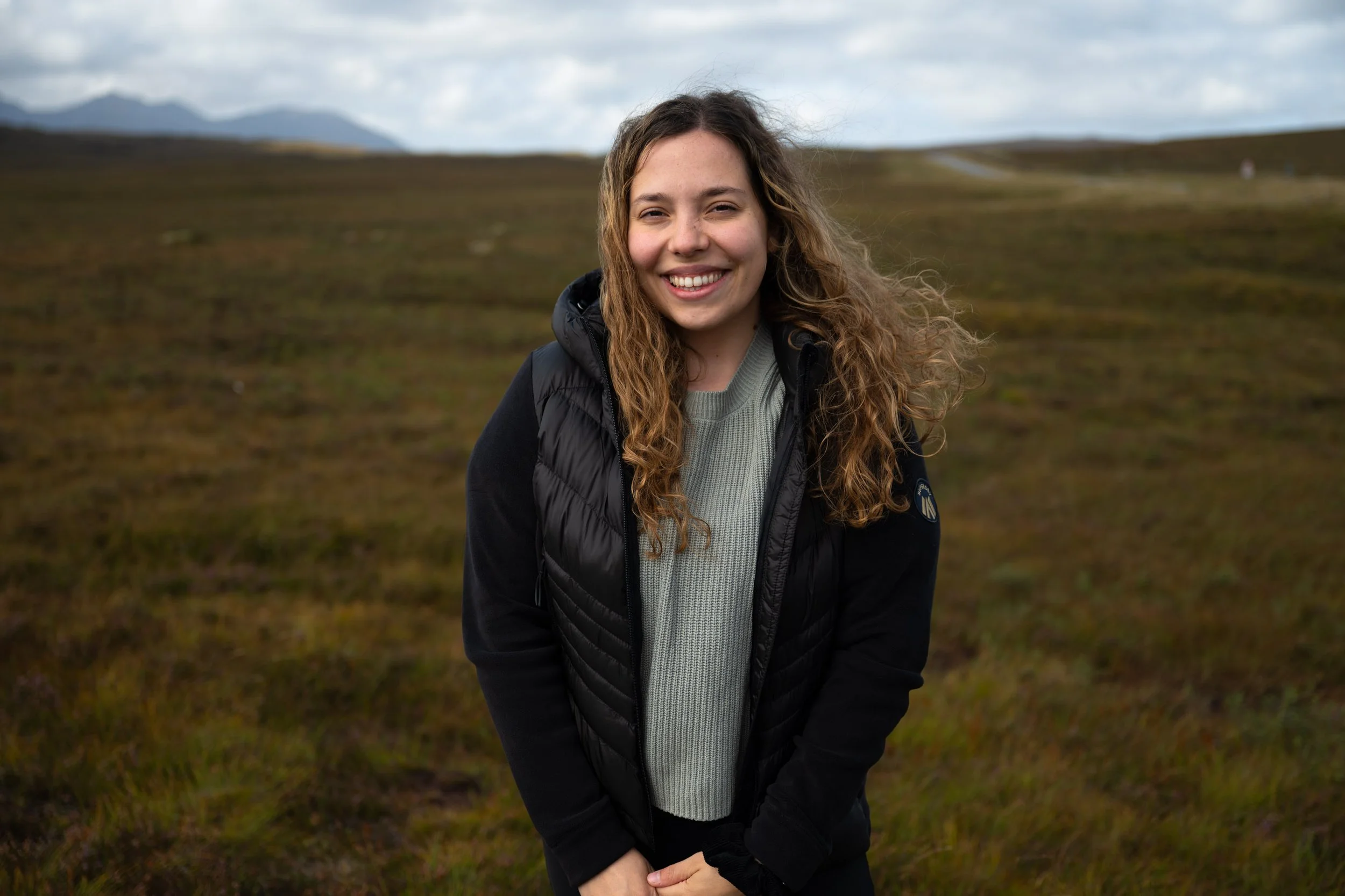 Junge Frau mit lang, lockigem Haar trägt schwarze Jacke und grauen Pullover, steht auf einer grünen Wiese bei bewölktem Himmel in Schottland. Dalia Schär.
