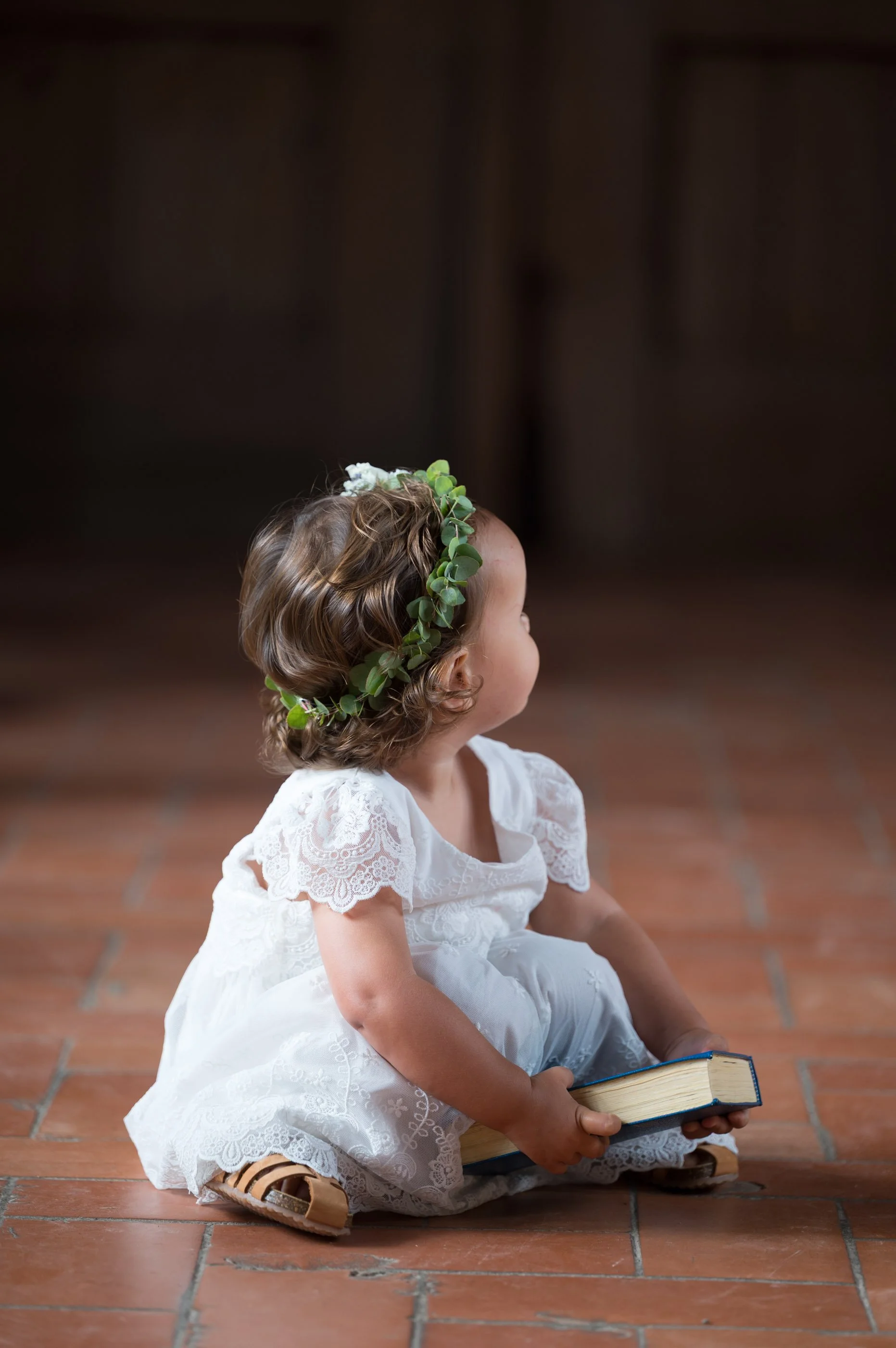 Kleines Mädchen im weißen Kleid mit Blumenkranz im Haar sitzt auf dem Boden, hält ein Buch in den Händen, auf einer Terakottabodenfliese.
