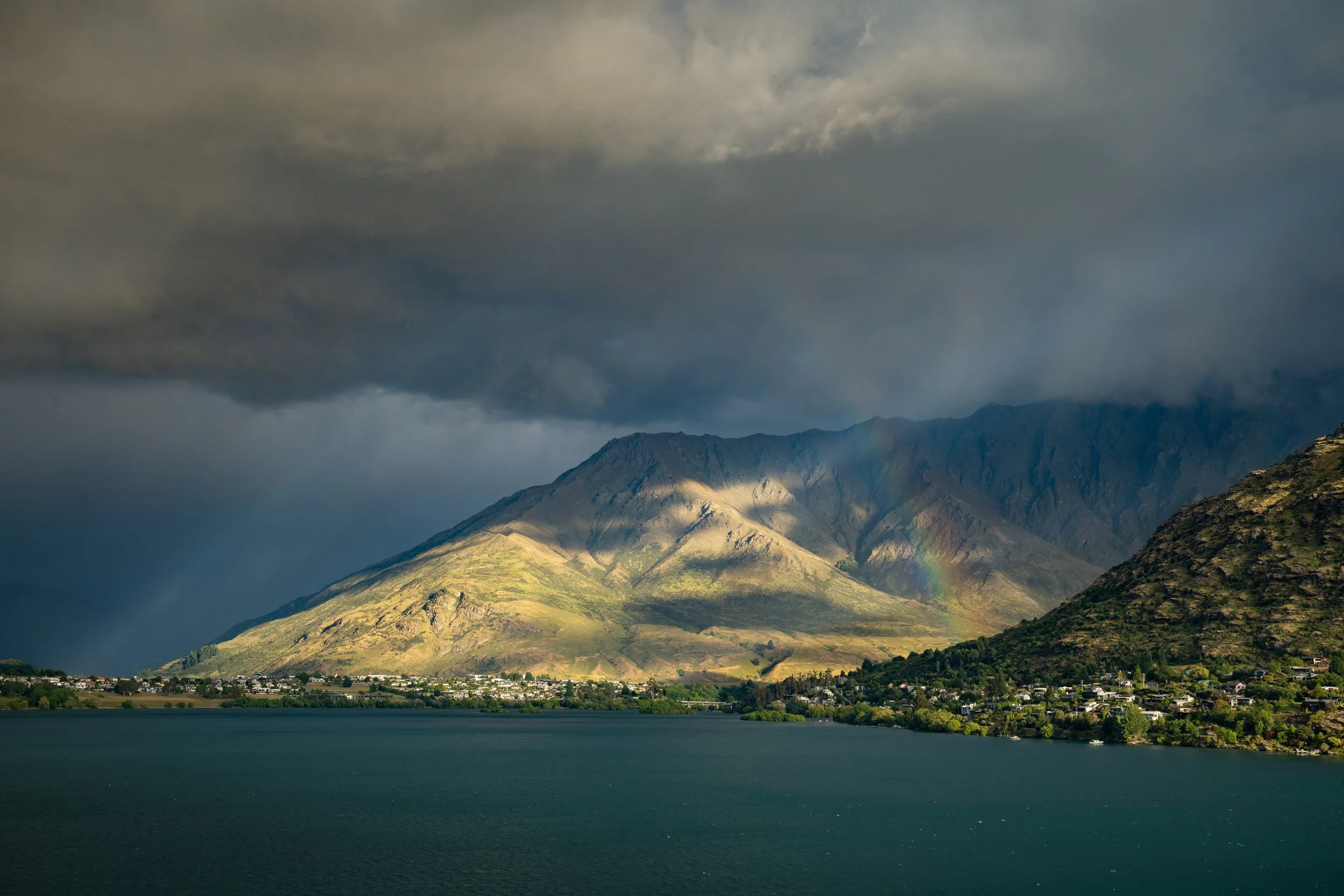 Berglandschaft mit dunklen Wolken, Wasser im Vordergrund, Sonnenlicht auf den Bergen und ein kleiner Regenbogen