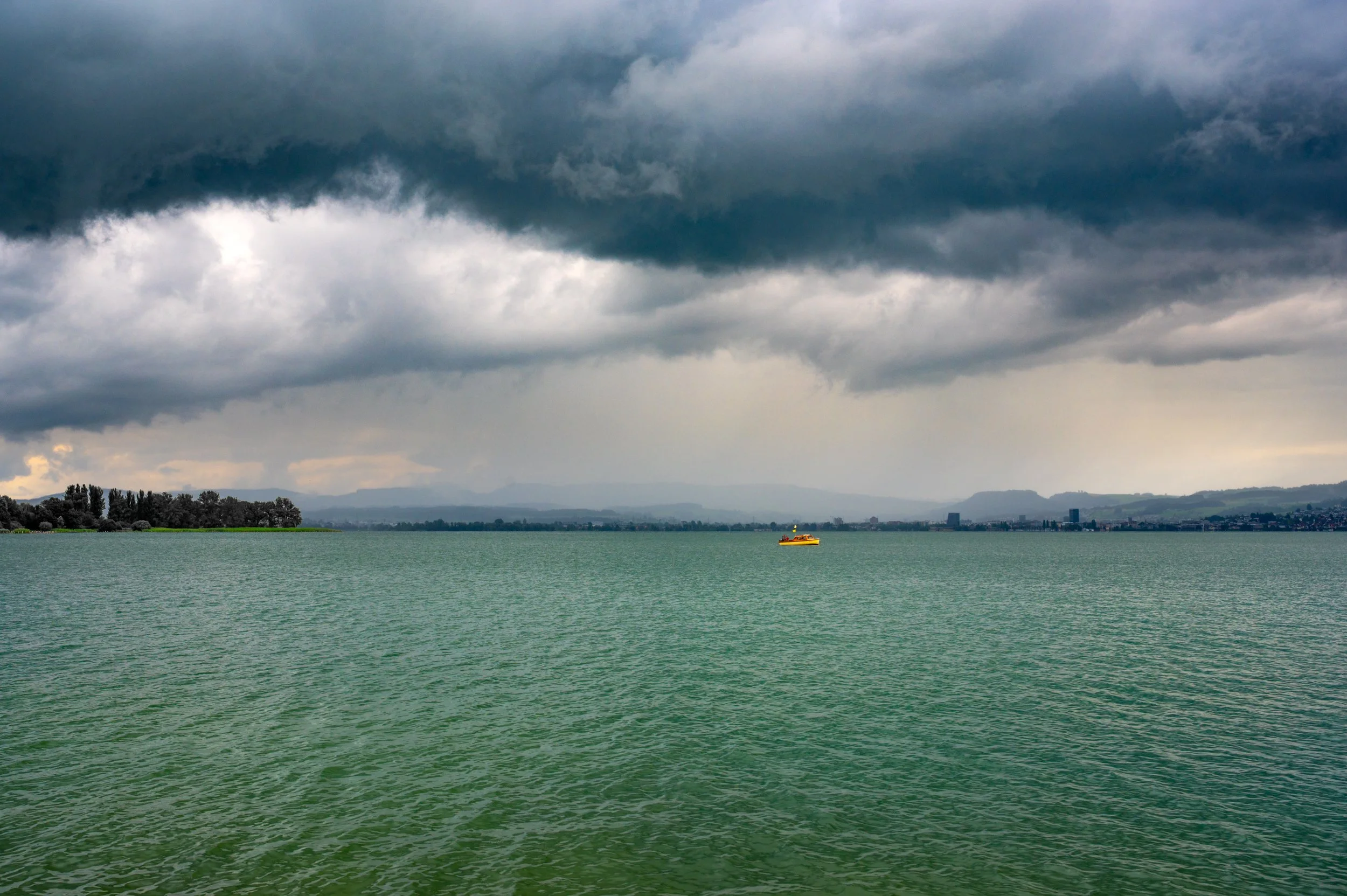 Blick auf einen großen See mit einer kleinen gelben Boot in der Mitte, dunklen Wolken am Himmel und einer Stadt im Hintergrund.