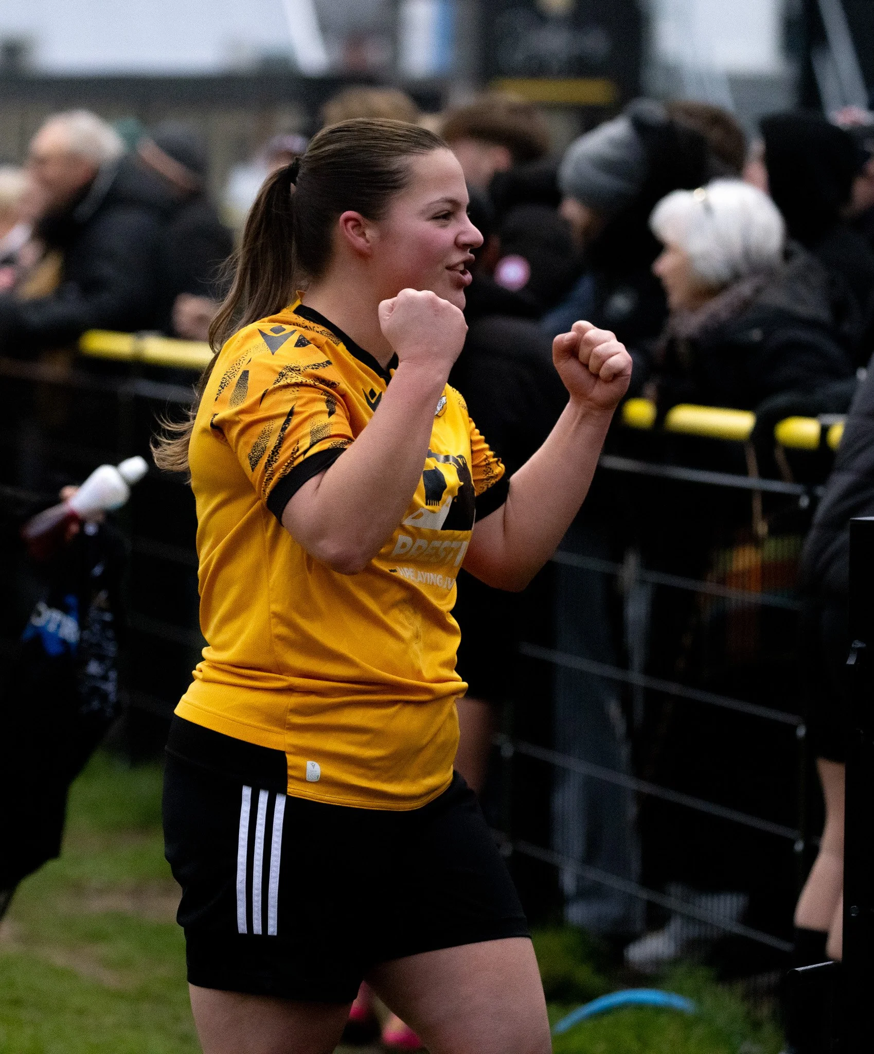 A young woman in a yellow sports jersey and black shorts is celebrating with clenched fists at a sports event, standing near a fence with spectators behind her.