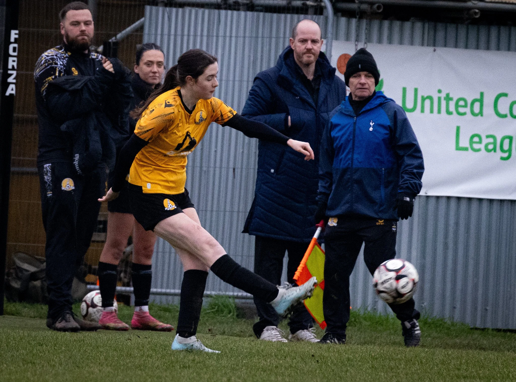 A woman soccer player in yellow jersey kicking a ball during a match. Two men and a woman in sportswear stand nearby, with one holding an assistant referee flag. A sign with green text reads 'United Co League' in the background.