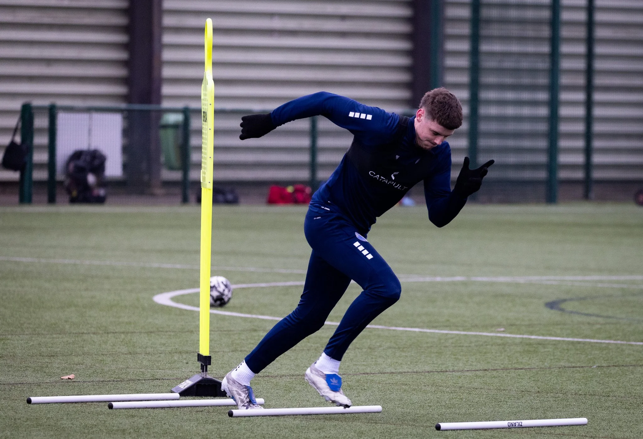 A male athlete in a navy training outfit runs during a training session on an outdoor sports field, passing a yellow hurdle, with soccer ball in the background.