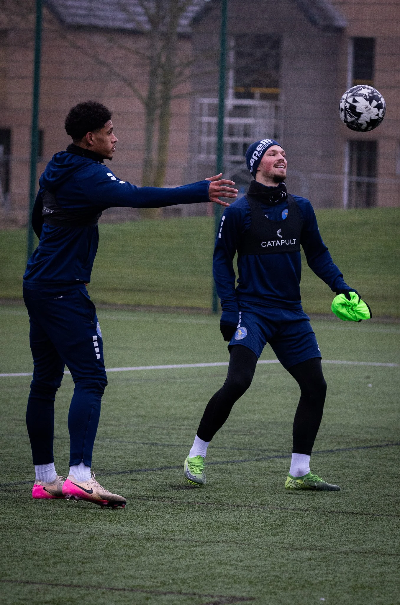 Two soccer players on a field, one passing the ball to the other, both wearing blue training gear, with a building and trees in the background.