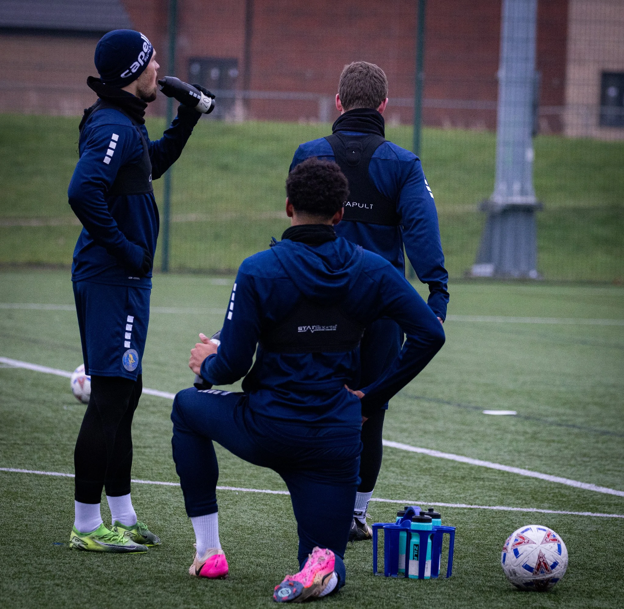 Three male soccer players in navy athletic wear on a field, one drinking from a water bottle, one kneeling, and one standing with his back to the camera, near soccer balls and water bottles.
