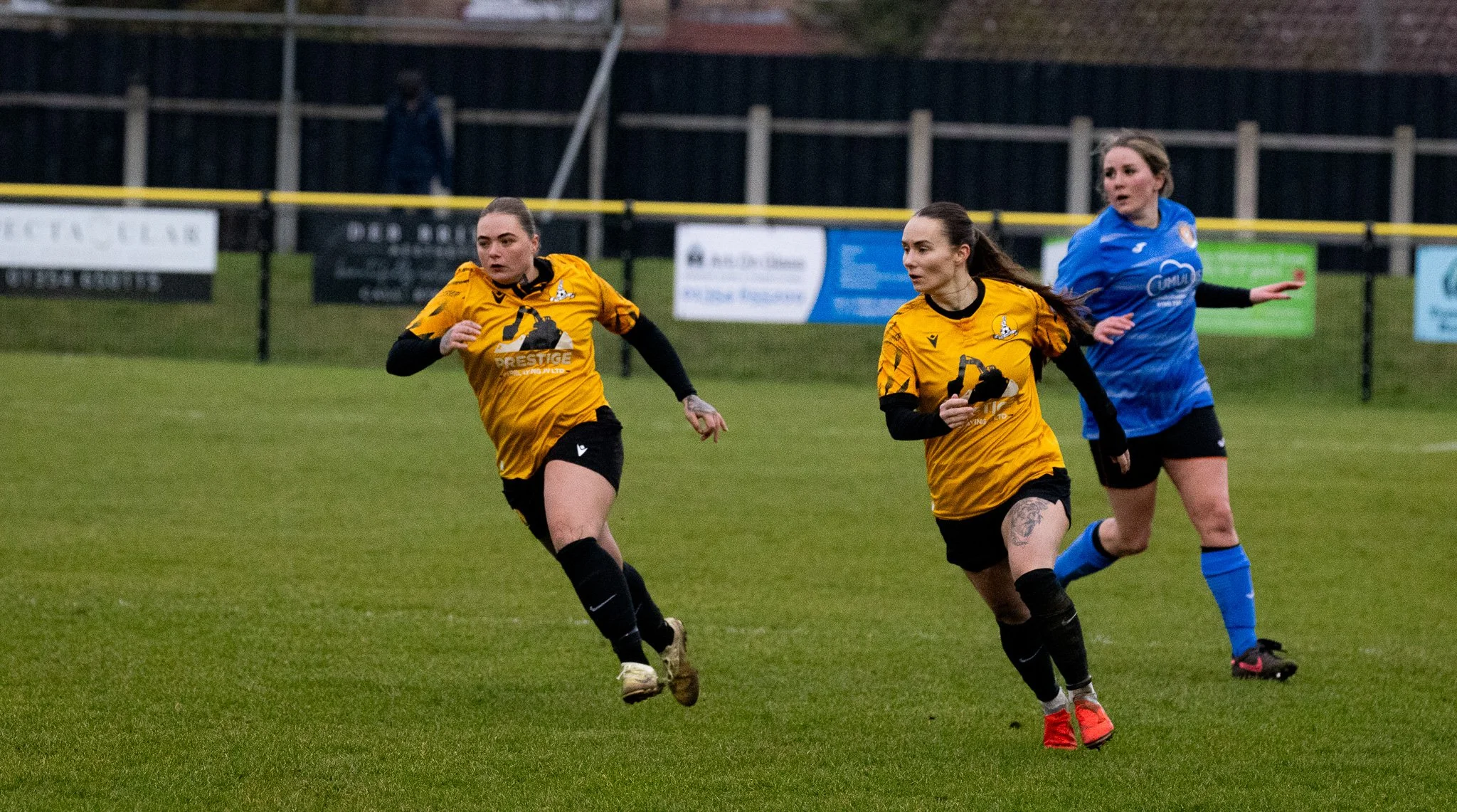 Three women playing soccer on a field, with two women in yellow jerseys and one woman in a blue jersey. Two women in yellow are running, and the woman in blue is behind them, all focused on the game.