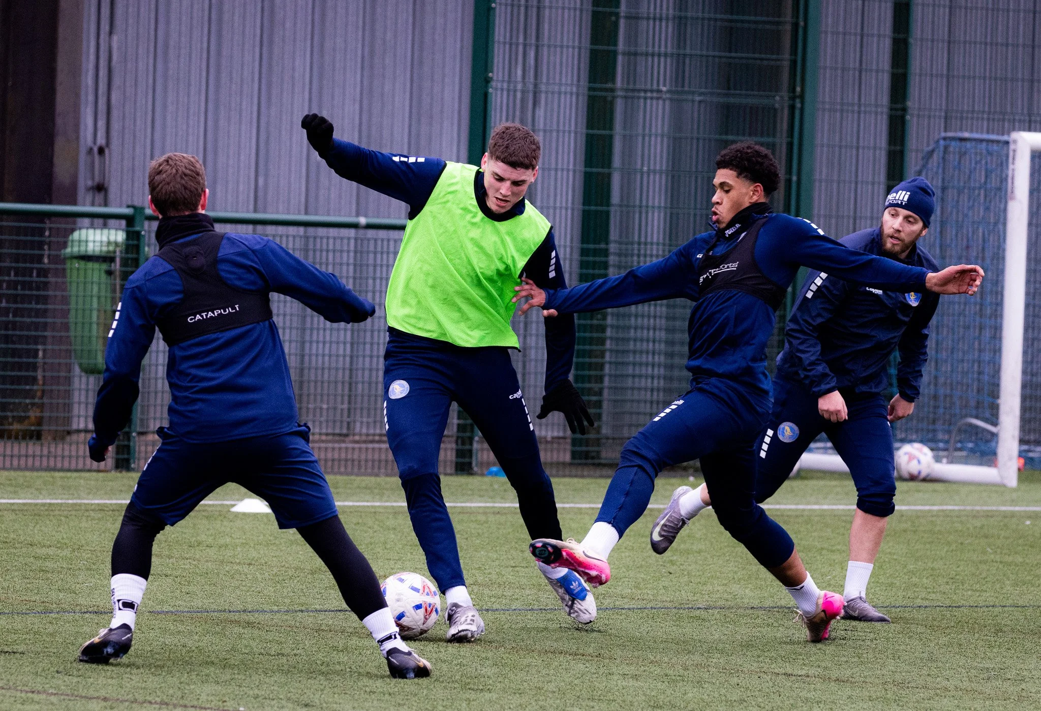 Four young men practicing soccer on an outdoor field, with one wearing a green training vest attempting to kick the ball while the others try to block. They are dressed in athletic gear, with a fence and goalpost in the background.
