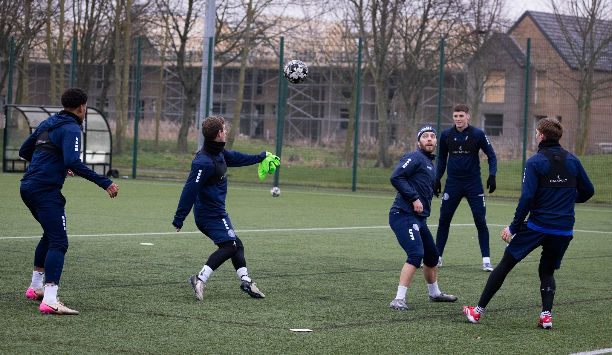 Group of five men playing soccer on an outdoor field, with a soccer ball in midair, trees, and buildings in the background during sunset.