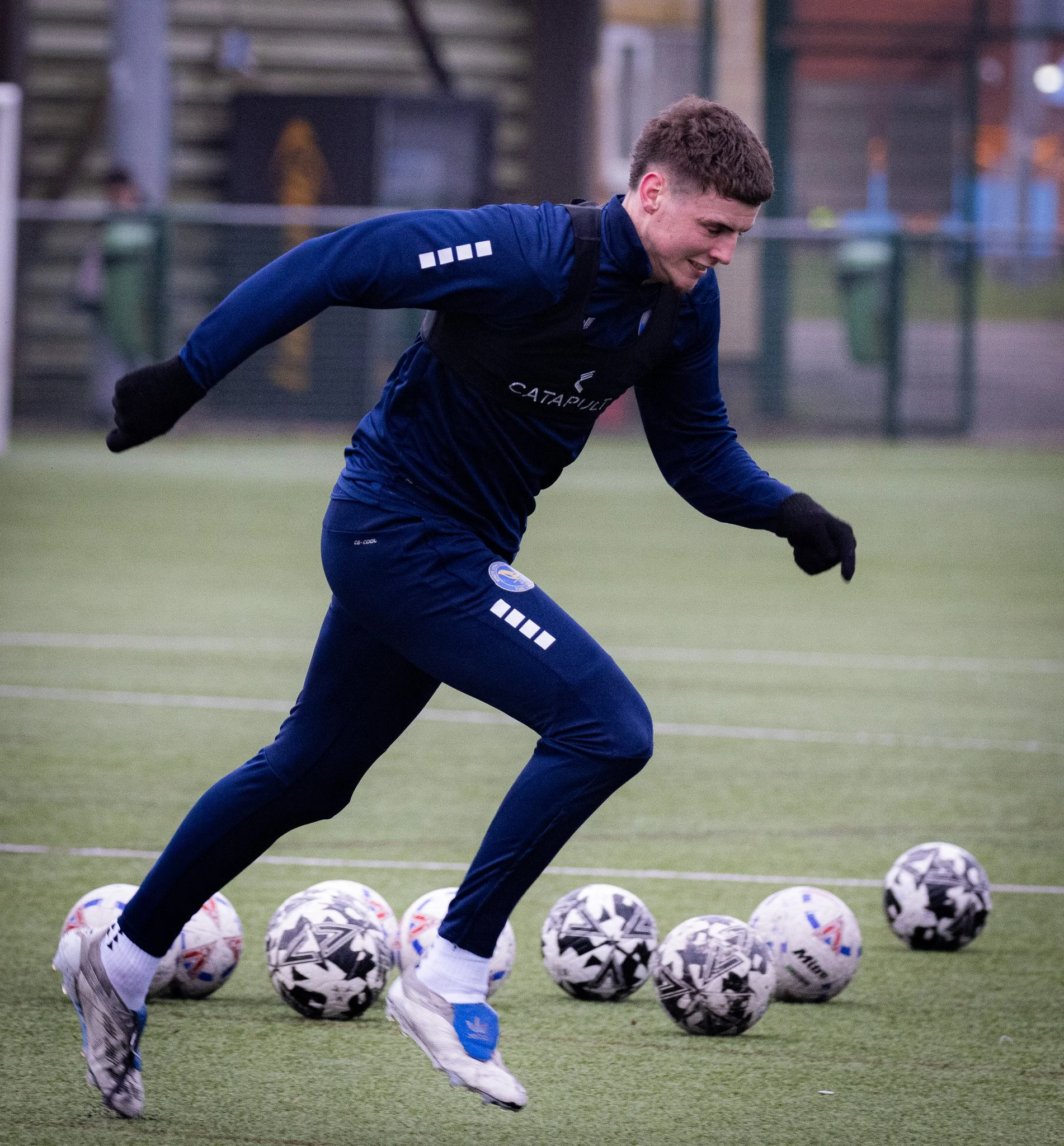 A male soccer player in blue athletic clothing and gloves practicing on a soccer field with multiple soccer balls arranged on the ground.