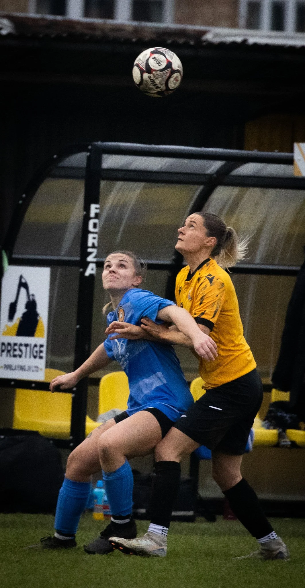 Two women are competing for the soccer ball during a game, with one wearing a yellow uniform and the other a blue uniform. The woman in yellow appears to be holding the woman in blue from behind as they both focus on the ball above them.