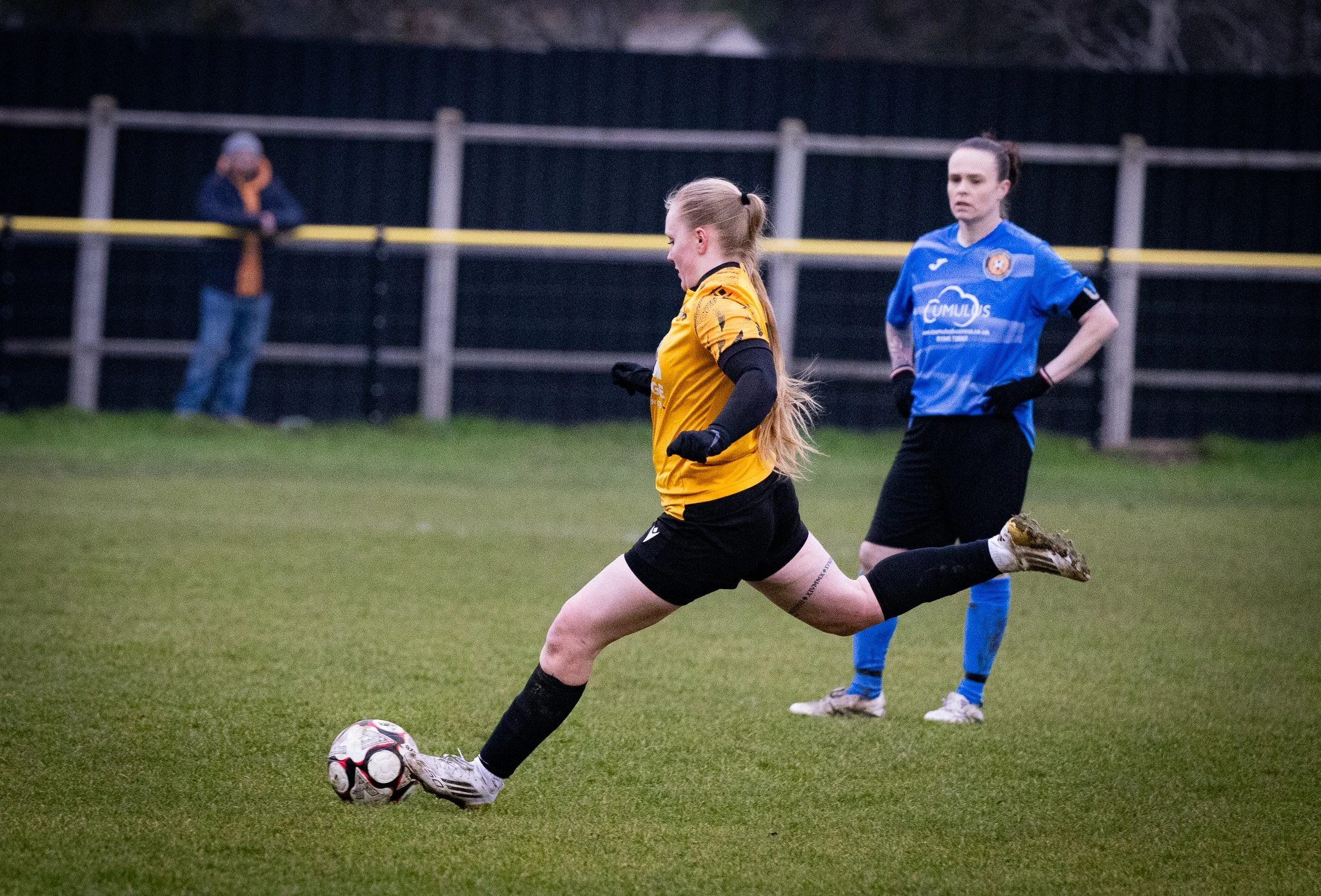 A female soccer player in a yellow jersey and black shorts kicking a soccer ball on a grassy field, with another female player in a blue jersey and black shorts standing in the background.