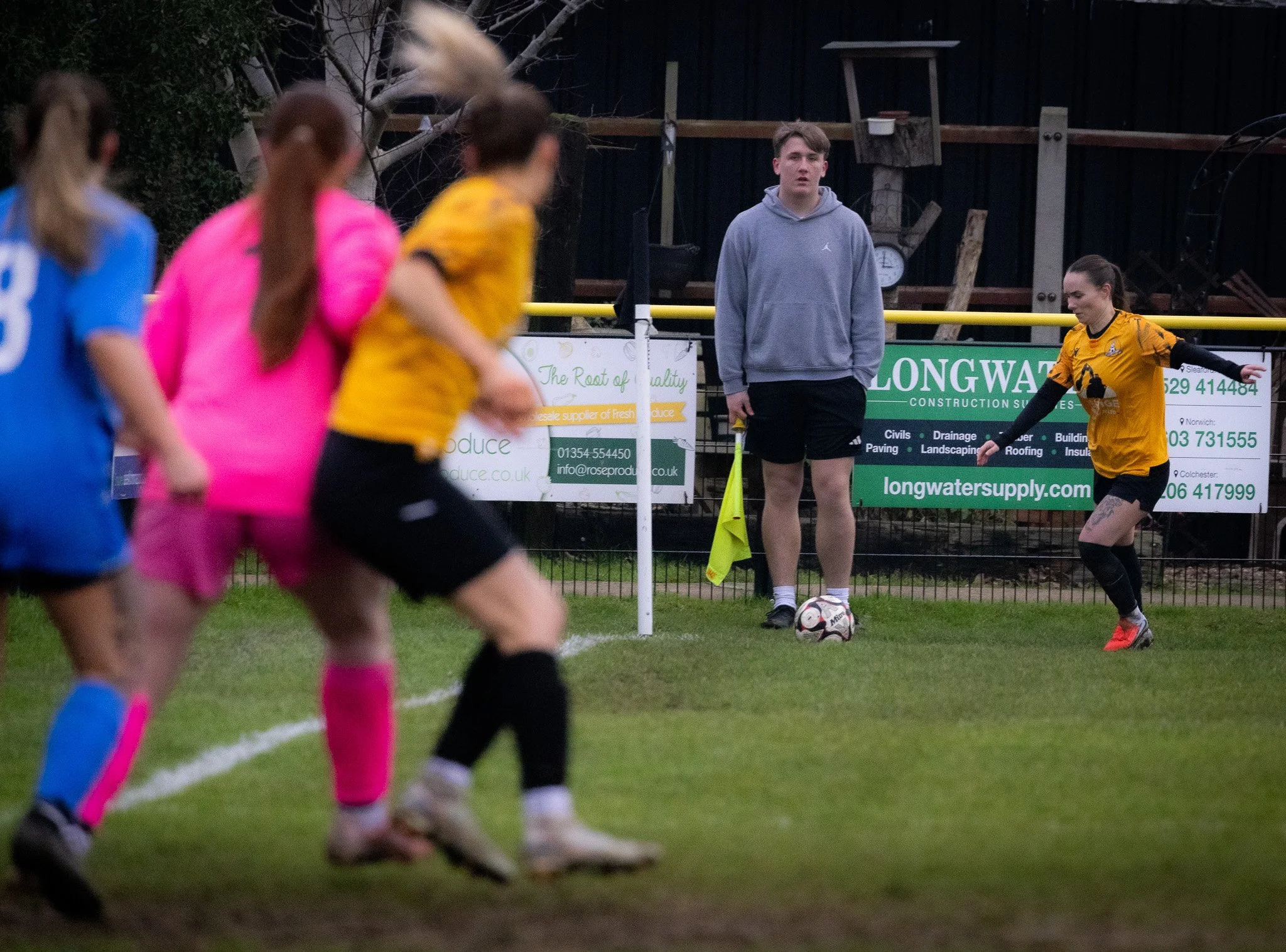 Women playing soccer on a grassy field, with one woman near a corner flag about to take a corner kick, and a young man standing near a soccer ball in the background