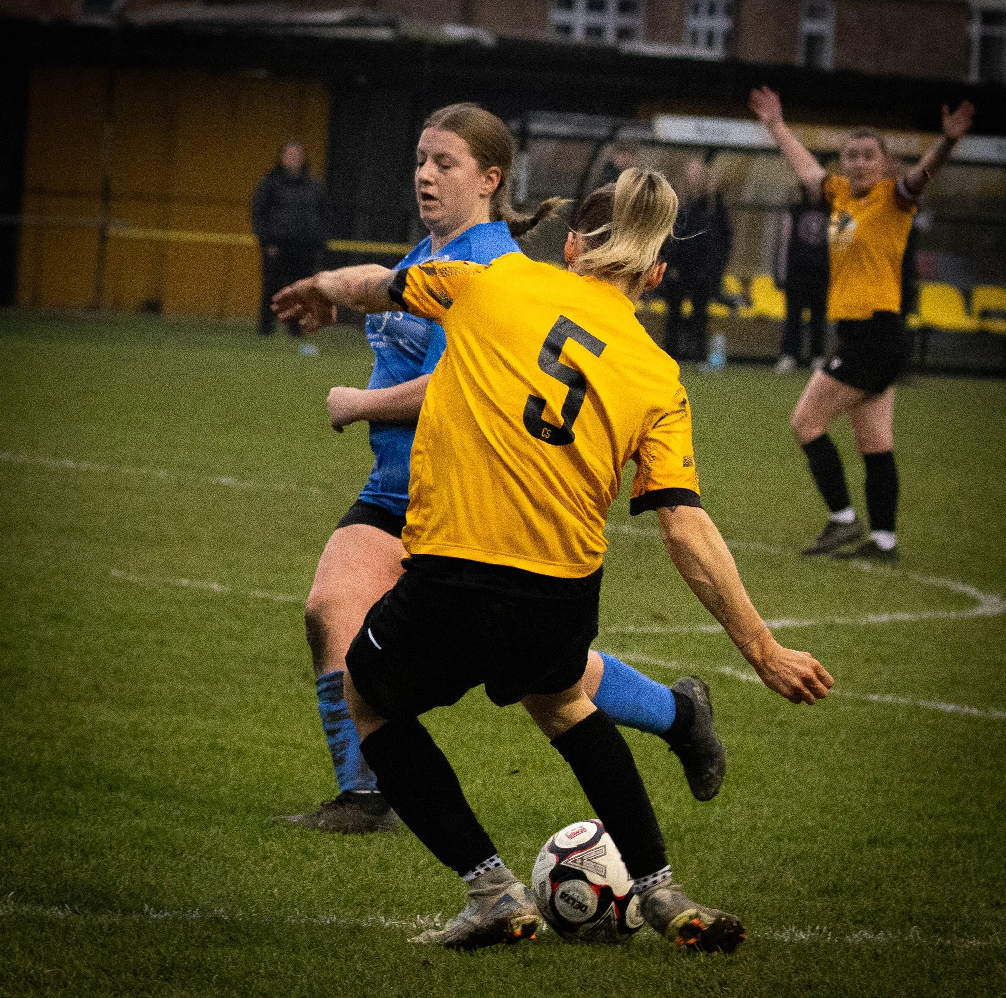 A soccer match with female players, one in a yellow uniform with the number 5, kicking the ball while another player in a blue uniform defends. Two more players are visible in the background on a grass field.