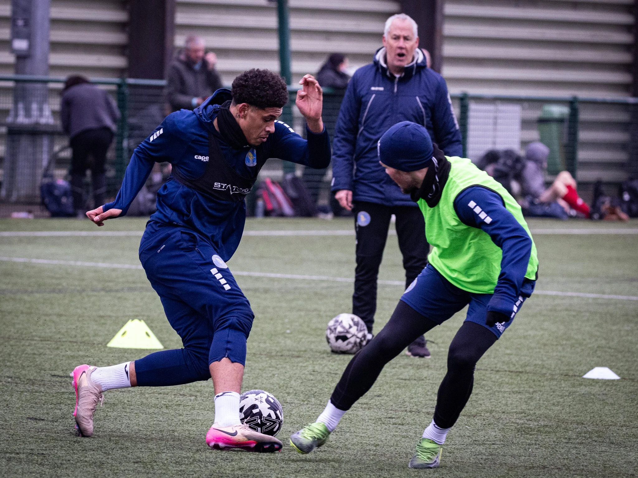 Two male soccer players practicing in a training field with a coach observing in the background.