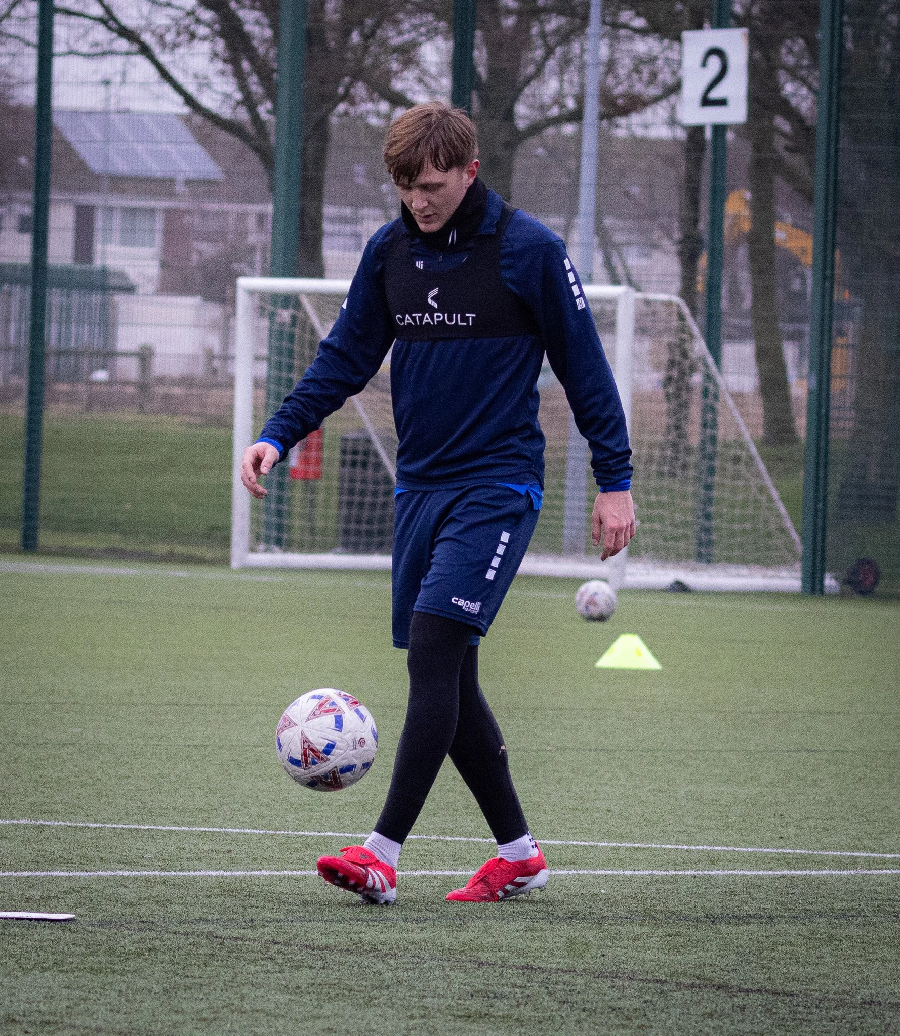 A young man in sportswear practicing soccer on a field, with soccer balls and training cones visible.