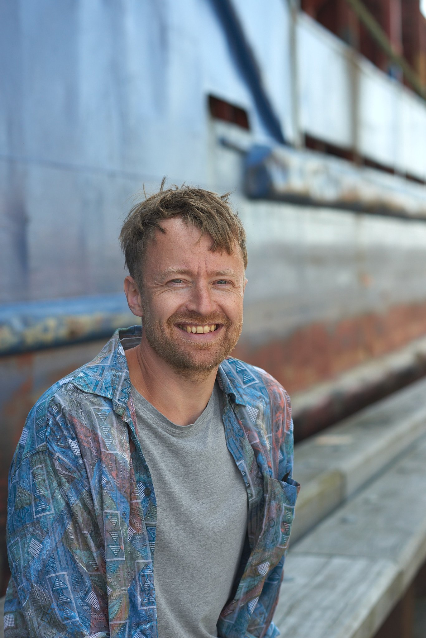 Smilende mand med lysthåret og skæg, siddende på en bænk udendørs, med en metallic båd i baggrunden.